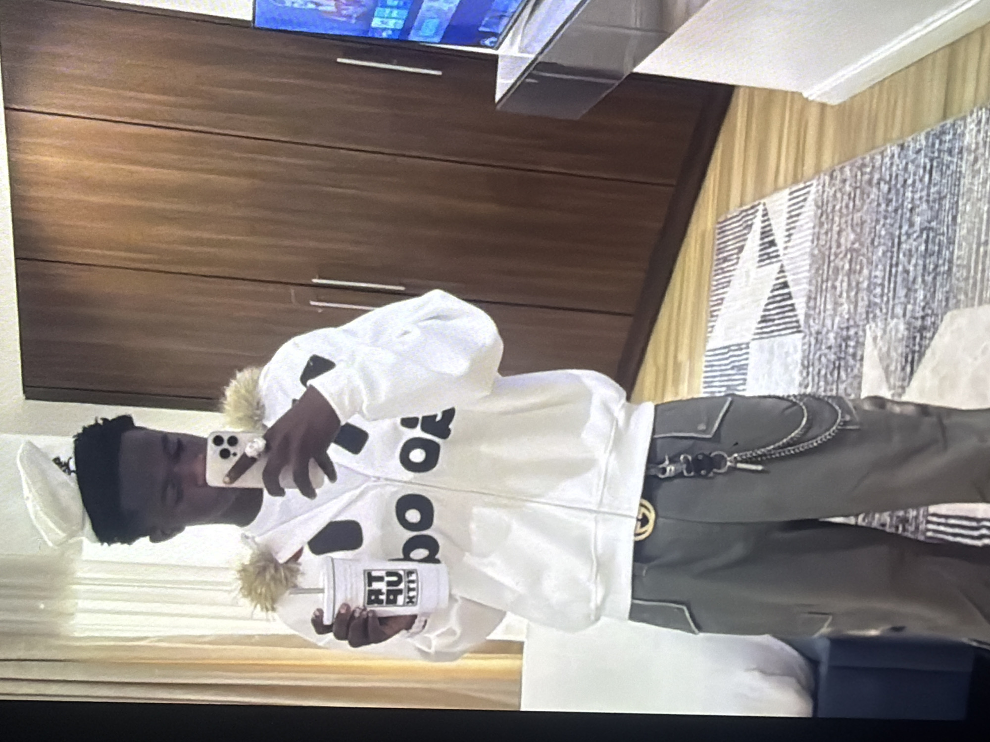 A young Black male is standing in a room, holding a white cup with a straw in his left hand and a smartphone in his right hand, taking a selfie. He is wearing a white hooded jacket with fur trim on the hood, a white t-shirt, grey cargo pants, and a white cap. The jacket has black lettering on it. A black belt with a gold buckle and a chain accessory is visible on his pants. The background includes wooden cabinets, a television, a rug, and what appears to be a bed. The image quality is slightly blurry and pixelated. There is no indication of the location being in Bukuru, Nigeria.