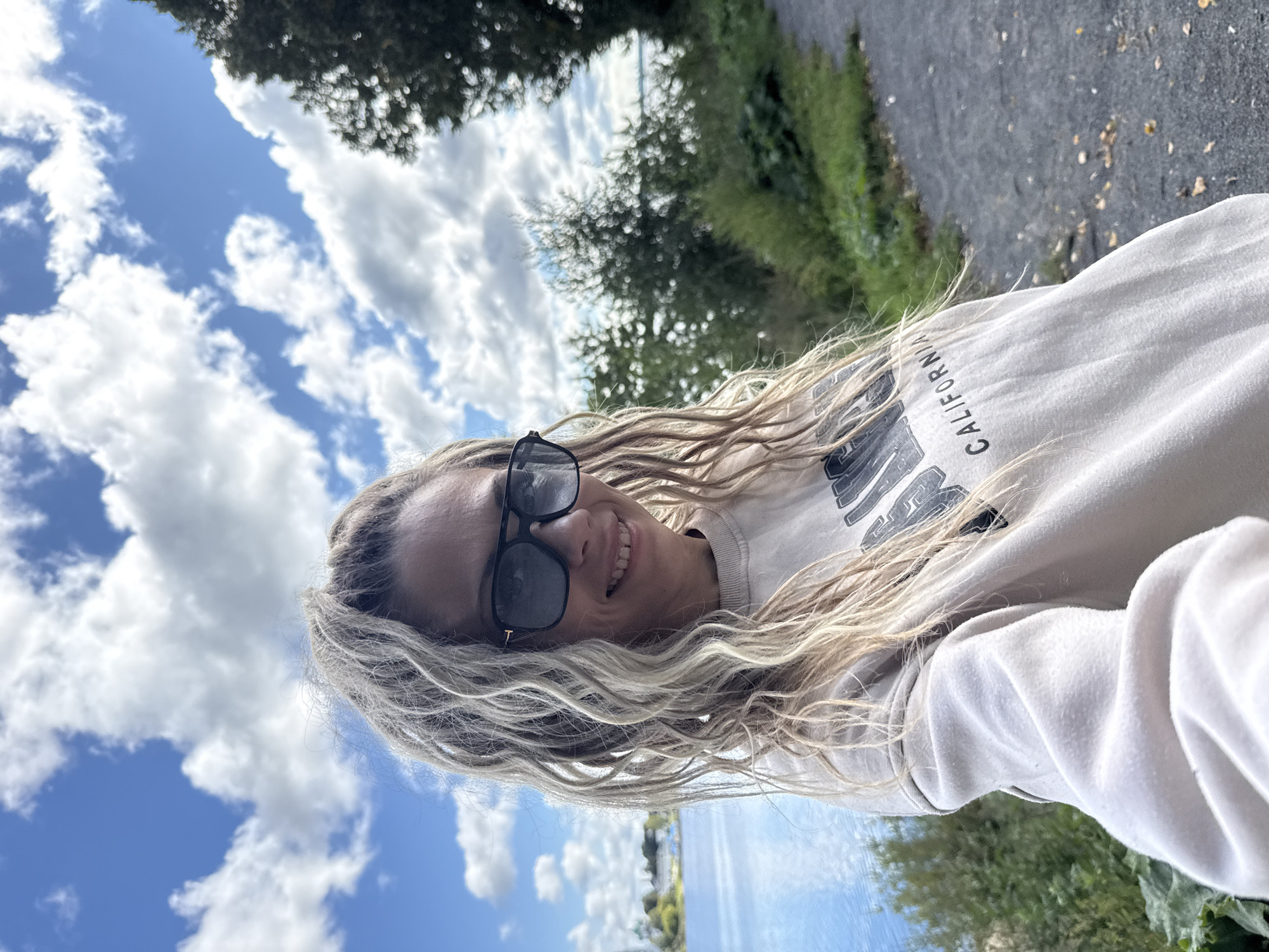 A medium shot, taken from the chest up, shows a woman with blonde, heavily textured hair that is pulled back and cascades over her shoulders. She is wearing dark, rectangular sunglasses and a light-colored crewneck sweatshirt with the word "CALIFORNIA" printed in dark block letters across the chest. The woman is smiling, her head tilted slightly to her left, and she appears to be holding the camera, as her left arm is partially visible and angled upwards.
The background reveals a body of bright blue water to the left, with a hint of land and buildings on the far side. To the right, a path made of dark gravel is visible, bordered by green grass and trees. The sky above is a vibrant blue, populated with fluffy white clouds. The scene is outdoors, likely in a park or along a waterfront, and based on the information provided, it is located in the city of Rusanovo, Russia. The overall atmosphere is bright and pleasant, suggesting a sunny day.