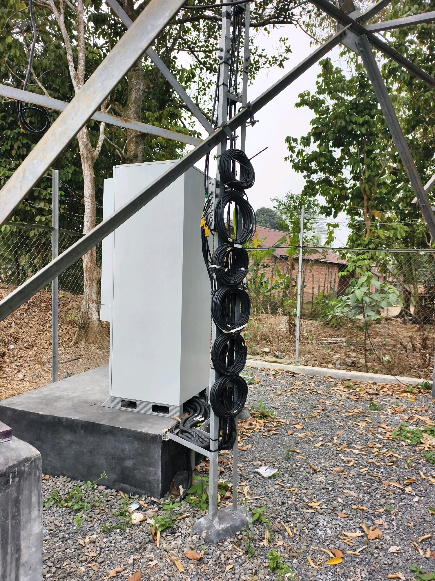 The image depicts a telecommunications tower setup in a semi-urban or rural environment, potentially in Simpang Jelutih, Indonesia. The scene is characterized by a white, rectangular equipment cabinet positioned on a concrete base, surrounded by gravel and dry leaves. A metal tower structure extends upwards, with multiple black coiled cables neatly bundled and attached to its vertical support. A chain-link fence runs along the perimeter, separating the site from an area with trees, vegetation, and what appear to be simple brick buildings with tiled roofs in the background. The lighting suggests an overcast or diffused daylight condition. No people are visible in the image.