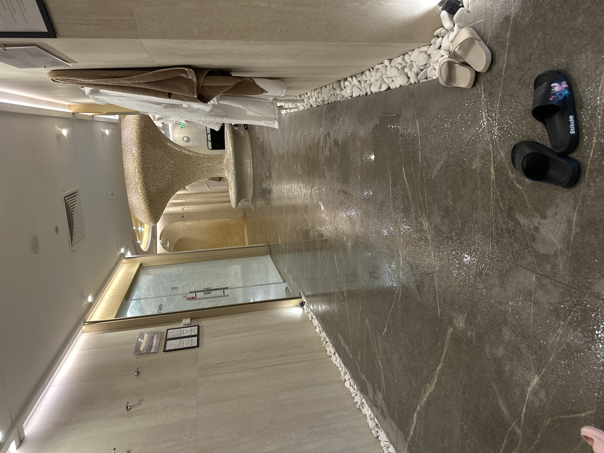 The image shows an interior space, possibly a spa or sauna area, with wet marble flooring and white pebble accents along the edges. A tanning bed is visible in the background, along with towels. In the foreground, two pairs of sandals are lying on the floor. The location is unknown, and there are no people present in the image.