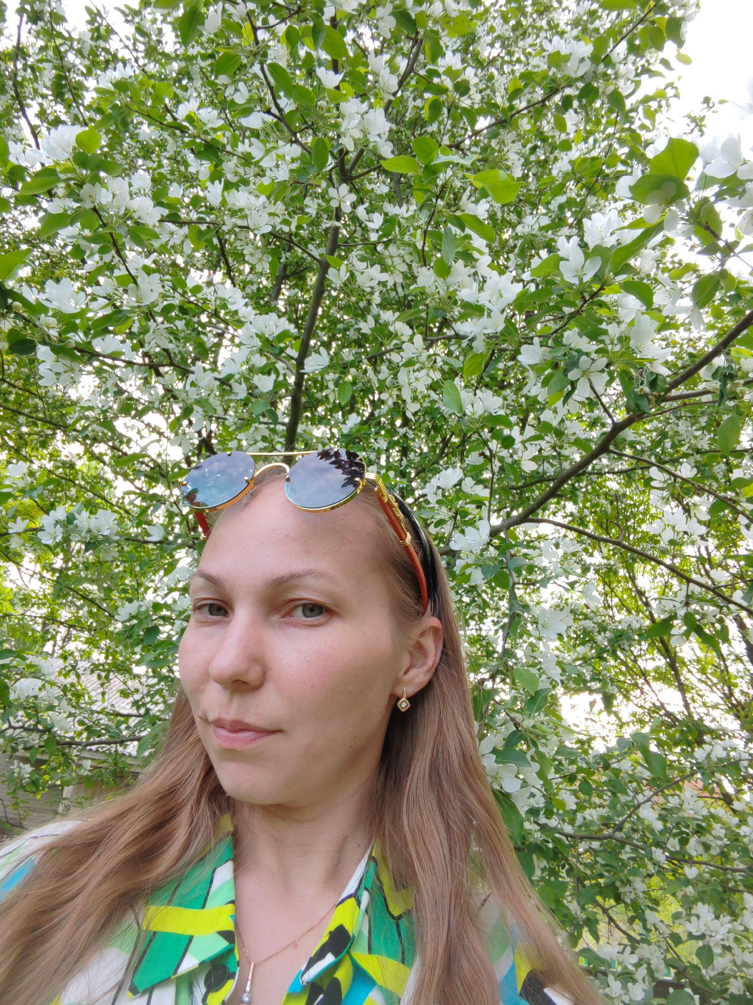 A young woman poses outdoors, framed by a flowering tree. She is wearing gold-rimmed sunglasses perched on her head, a green and white patterned shirt, and a delicate gold necklace. She has long, light brown hair and wears small gold earrings. The location appears to be a park or garden in Чухлэм, Russia, as the flowering tree is a common feature of the region's landscapes. The scene is bright and sunny, suggesting it is daytime.