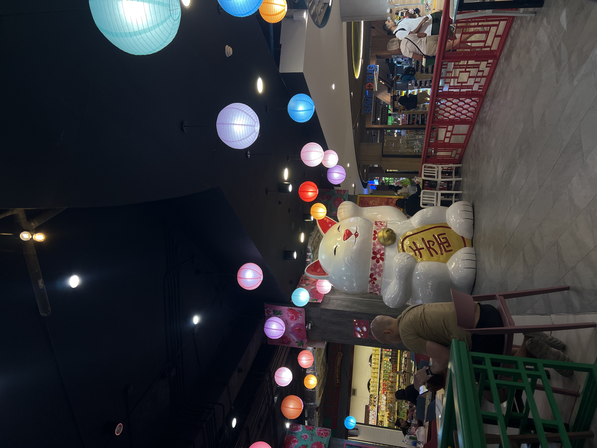 The image shows an indoor scene, possibly a restaurant or food court, located in an unknown city. The dominant feature is a large, white, ceramic maneki-neko (lucky cat) statue with a golden coin-shaped decoration that has yellow writing on it. Above the cat are colorful hanging lanterns, illuminated by lights. The ceiling is primarily black.
In the background, there are food and drink vendors behind the cat, showing various merchandise. Near the cat's base and next to a green chair, a person is sitting and appearing to be looking at a phone. A red metal fence-like structure is also visible. In the distance, there are other people, seated or standing, in the space. The general atmosphere suggests a public eating or shopping area.