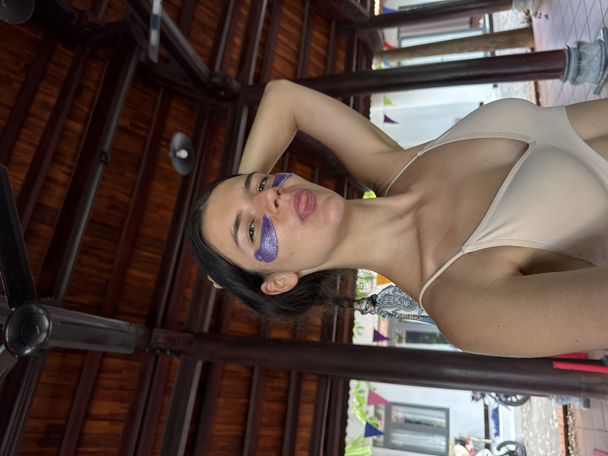 This is a medium shot, taken at eye level, of a young woman with dark hair and fair skin, lying on her back and looking at the camera. She is wearing a light-colored tank top and has purple under-eye masks on. She has her left arm bent behind her head, and her right arm is out of frame. Her tongue is sticking out slightly. The background is a ceiling with wooden beams, and there is a ceiling fan visible. Colorful triangular flags are hanging in the distance, suggesting a festive or outdoor setting. The lighting appears to be natural, possibly from an overhead source, and there are no strong shadows, indicating it may be daytime. The overall mood seems playful and relaxed. The location context provided is Mũi Né, Vietnam. No visible text is present in the image.
