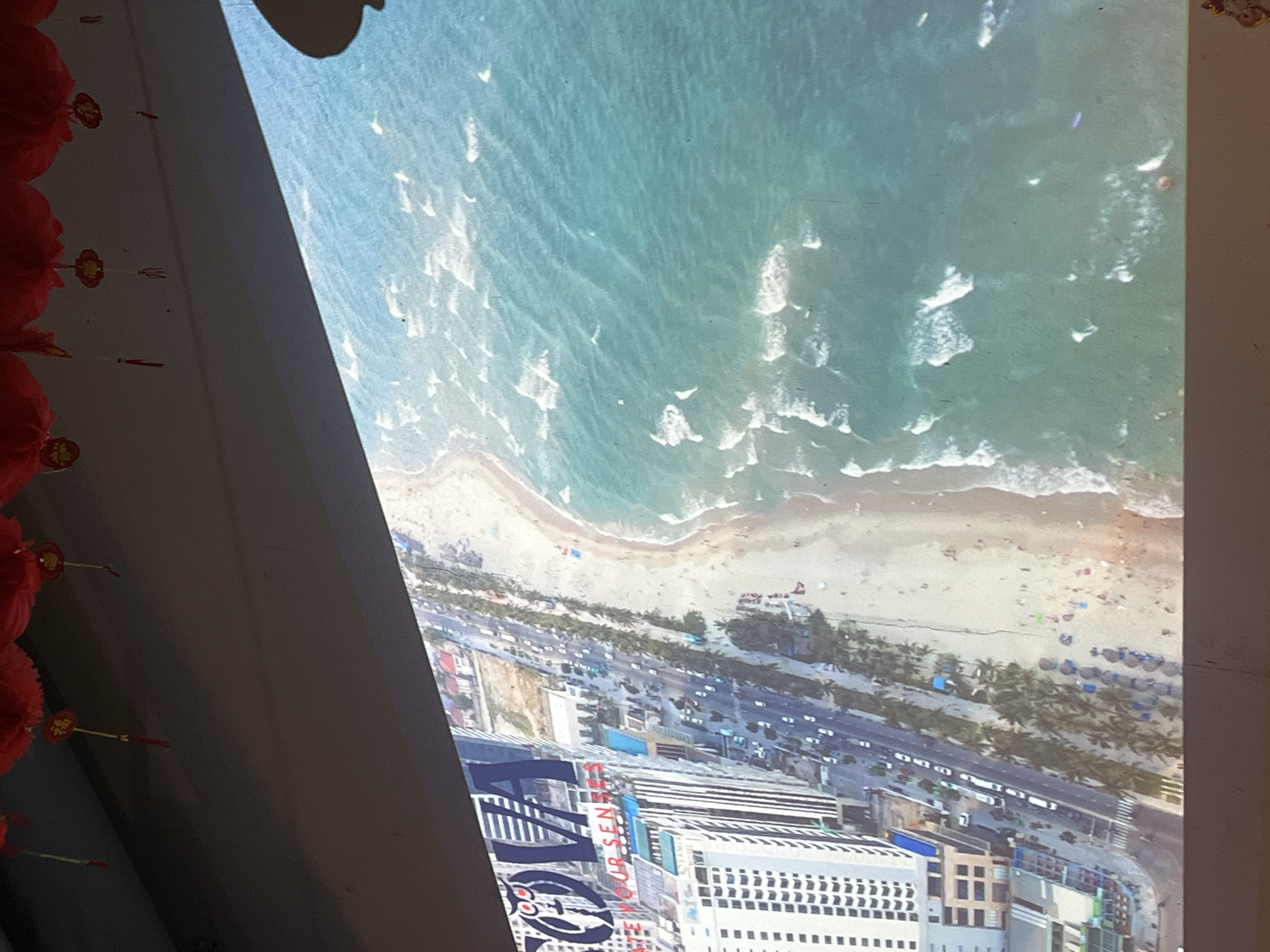 The image shows an aerial view of a coastal area in Đà Nẵng, Vietnam. The scene includes the shoreline with waves breaking onto the beach, a sandy beach with people and beach umbrellas visible, a coastal road with traffic, and buildings of varying heights in the city. The water is turquoise. There are palm trees. Buildings include a hotel with the words "NOVOTEL" visible. Red lanterns are hanging nearby.