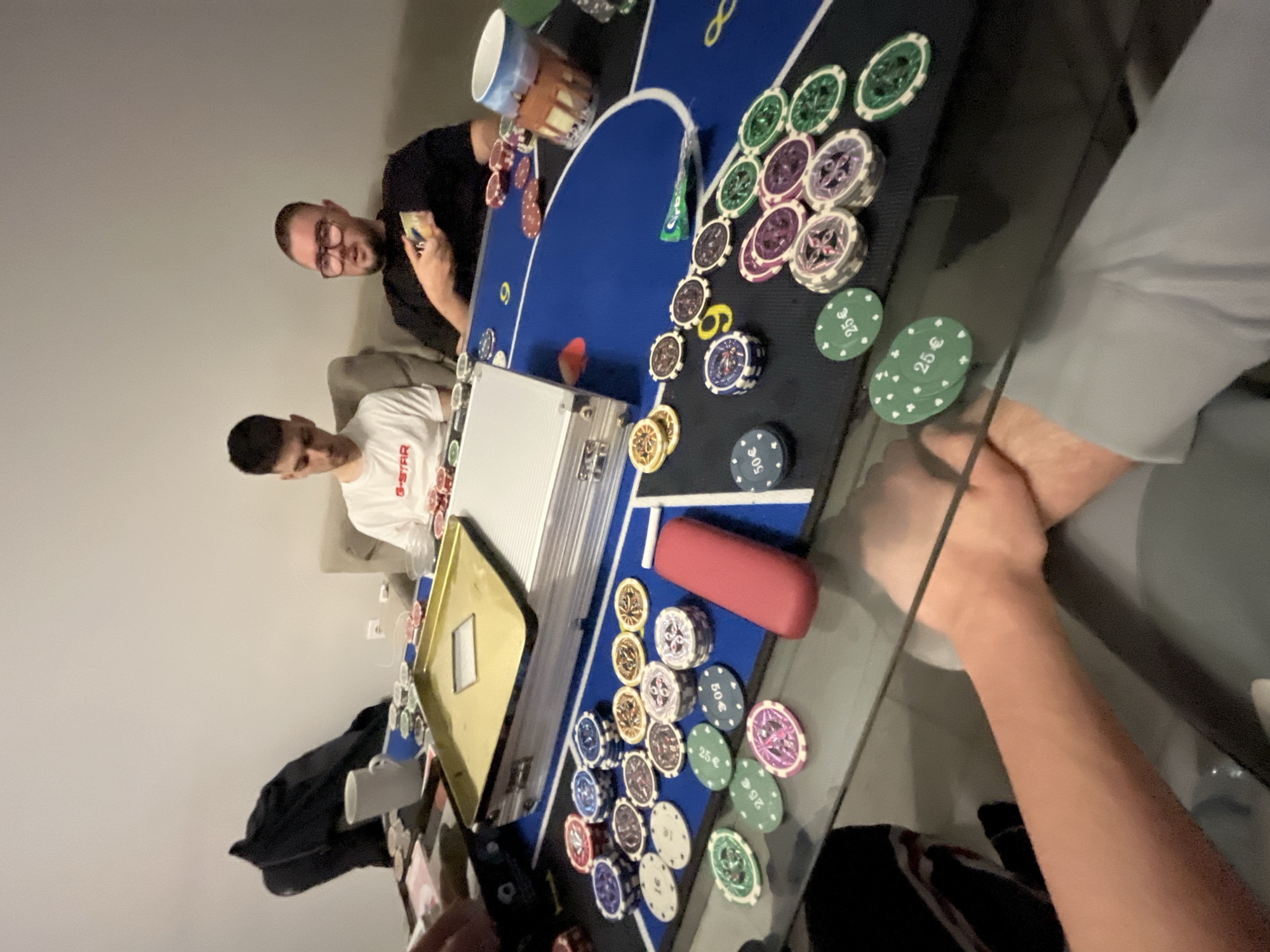 The image captures an indoor scene, rotated ninety degrees clockwise, depicting a casual poker game in progress. Three individuals are visible around a table with a blue and black poker mat.
In the upper portion of the frame (left after rotation), a man with glasses wearing a dark shirt is seated, intently holding a hand of playing cards. Further down the table (center after rotation), another man in a white t-shirt with "G-STAR" printed on it is also seated, looking down towards the table. The arm and hands of a third person are visible in the foreground on the right side of the frame, resting on the table.
The table is covered with a blue and black poker mat, featuring various outlines for cards and chips. Numerous poker chips in different colors—green, purple, white, blue, red, and gold—are scattered and stacked across the mat. Some green chips clearly display "25 €" and some blue chips show "50 €" as their denomination. A silver poker chip case is open on the table, along with a golden tray or lid next to it. A red rectangular object, possibly a card protector or chip holder, is also on the mat. A white ceramic mug and another patterned cup are visible beside the players.
The background is a plain light-colored wall, suggesting a home or casual setting. The artificial lighting indicates it might be evening or night. The people appear focused on the game, creating an atmosphere of friendly competition.