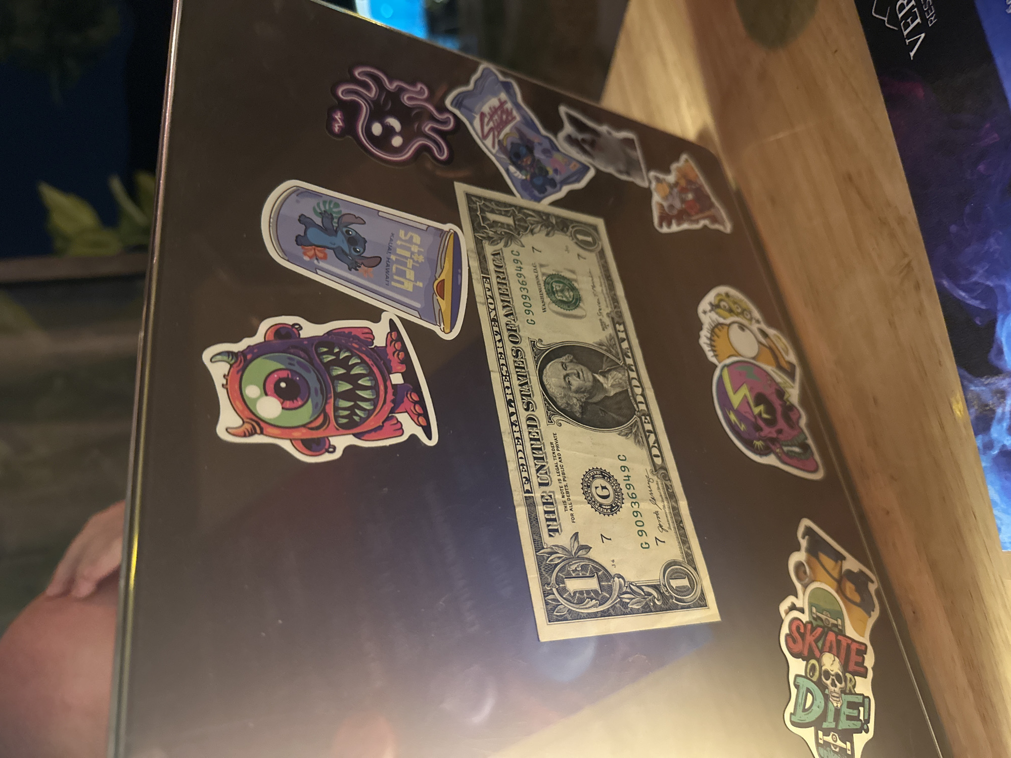 A photograph captures a laptop adorned with assorted stickers and a US one-dollar bill. Stickers include cartoonish images of a monster, a skull with lightning, and characters from the Disney movie "Lilo and Stitch". The dollar bill is placed diagonally across the laptop's surface. The location appears to be indoors on a wooden table, with a book and greenery visible in the background. The location within Bac Nha Trang Ward, Vietnam, cannot be confirmed from this image.