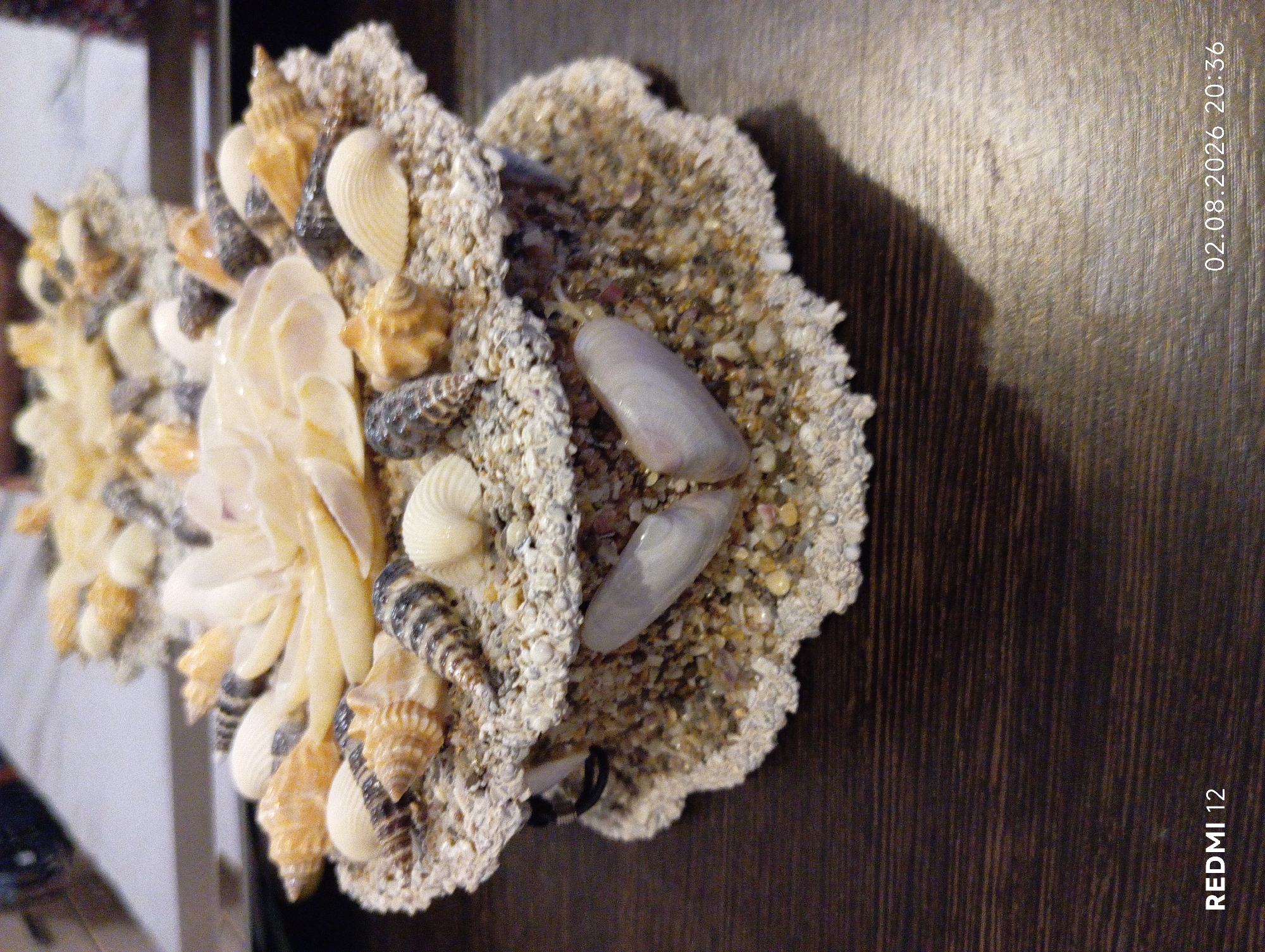 The image shows a close-up view of a decorative item made of seashells and sand, placed on a dark wooden surface. The item is shaped like a layered shell or a tiered cake, with various types of seashells embedded in sand. The bottom layer appears to be a rough, irregular shape, while the top layer is more circular and adorned with larger, lighter-colored shells in the center, resembling petals. Smaller, darker, and patterned seashells are arranged around the edges of both layers. Two elongated, pale shells are visible on the front of the bottom layer. The lighting casts a shadow on the wooden surface. The bottom right corner of the image contains a watermark reading "02.08.2026 20:36" and "REDMI 12". There is no indication of the city of Oryol, Russia, or any people in the image.