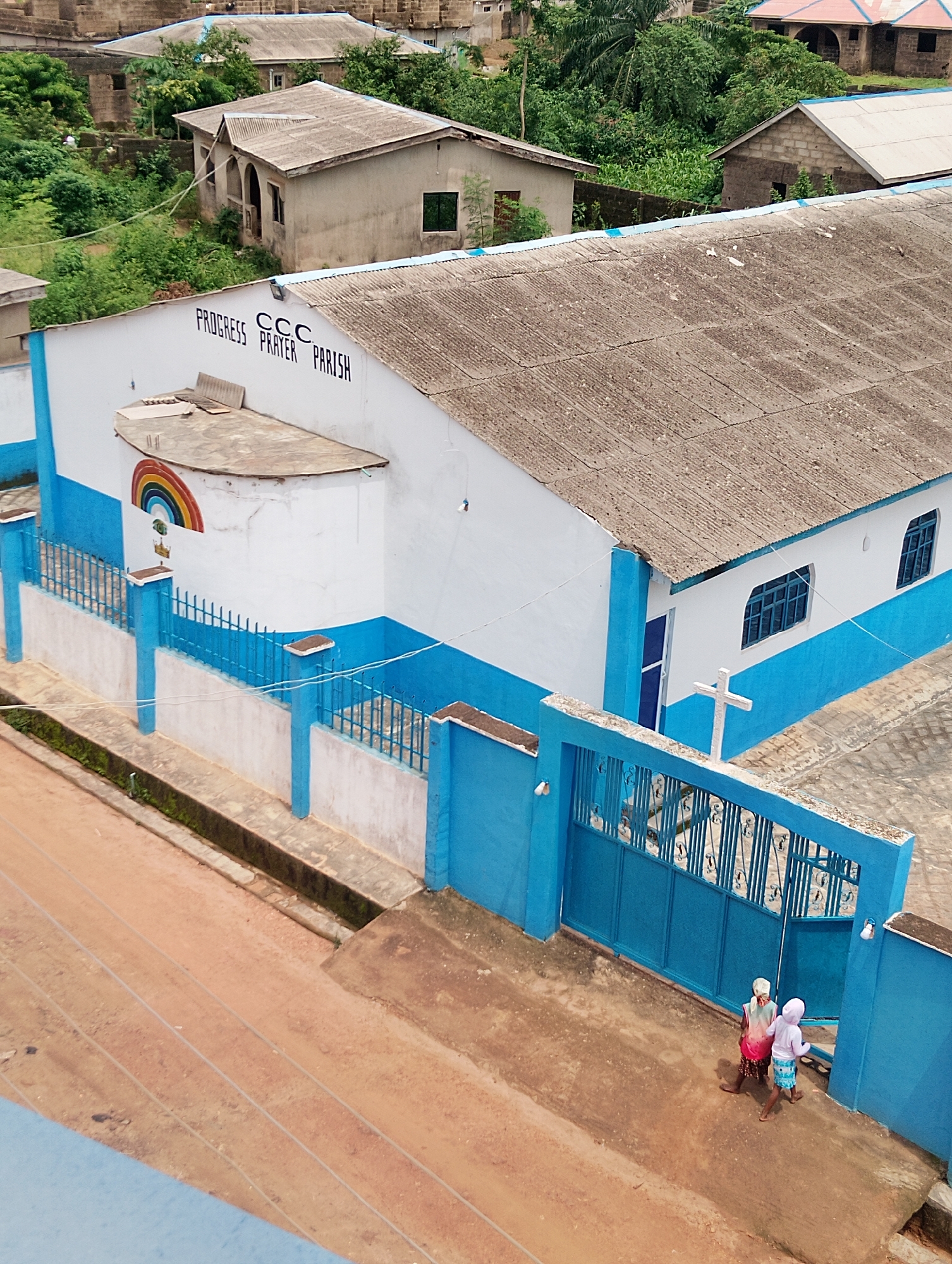 An aerial shot captures the exterior of the CCC Prayer Parish in Agege, Nigeria. The building features white walls with a prominent blue stripe along the lower half and a corrugated roof. The church is partially enclosed by a blue metal fence, with a matching gate. A white cross is visible on a concrete pillar near the entrance. A painted rainbow mural adorns the side of the building. Two individuals, one in a pink and patterned outfit and the other in a light pink hooded garment and blue shorts, are walking towards the blue gate. The surrounding environment includes other buildings, dense green foliage, and a reddish-brown dirt path.