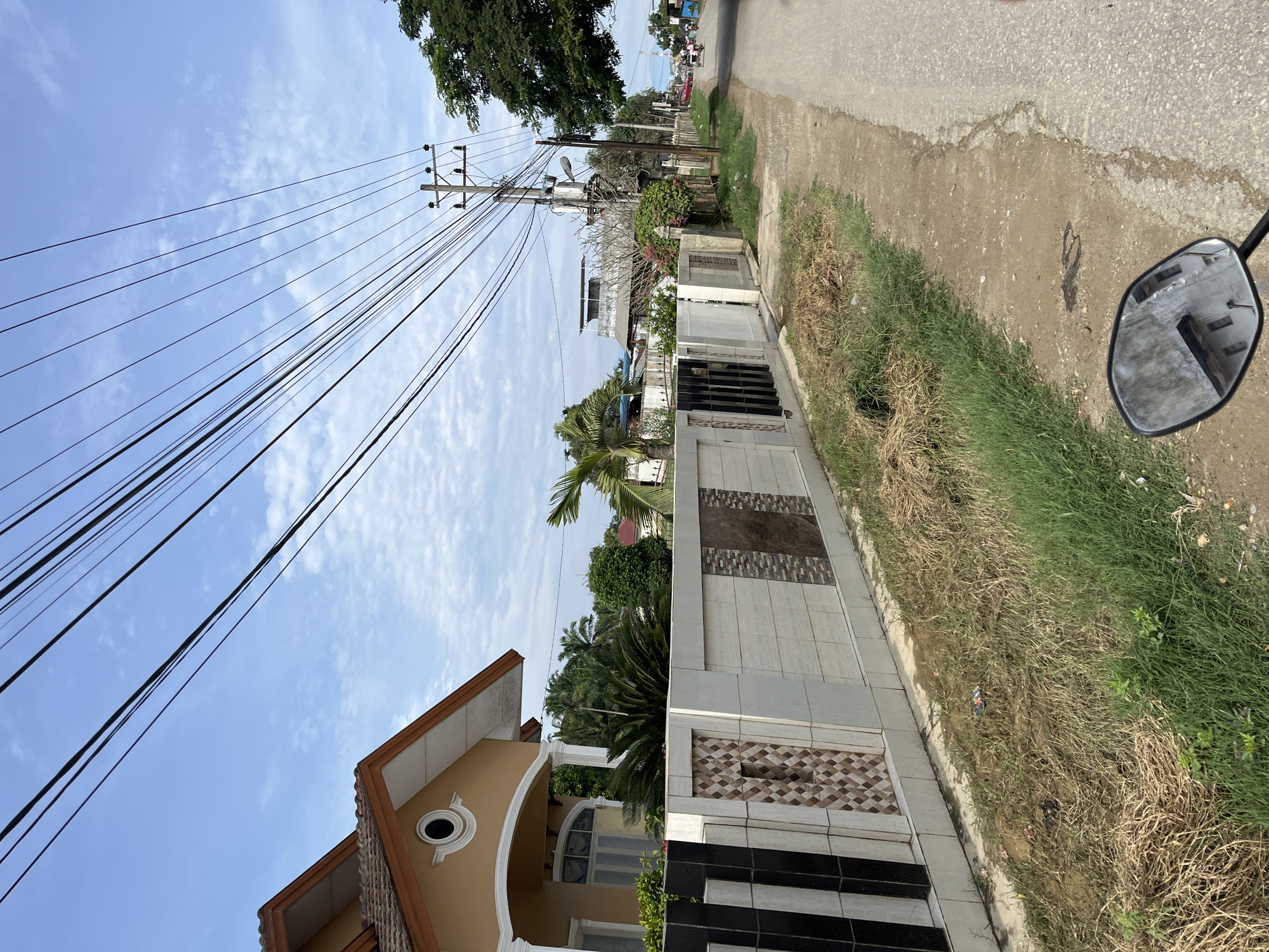 The image captures a street scene in Landuh, Indonesia, on a clear day with scattered clouds. On the left, a section of a cream-colored house with an orange-tiled roof is visible, featuring a decorative circular window. A white tiled fence with black and brown patterned accents runs along the property line, separating it from the street. Beyond the fence, palm trees and other greenery are present.
The street itself is paved, with a dusty shoulder on the left. A prominent utility pole with electrical wires crisscrossing the sky stands near the center of the frame. Further down the street, several more houses and trees can be seen, along with other utility poles. A few vehicles, including a tricycle and a motorcycle, are visible in the distance on the right side of the road, suggesting a modest level of activity. The foreground shows the side mirror of a motorcycle, reflecting the environment. The overall impression is of a residential area in a tropical climate.