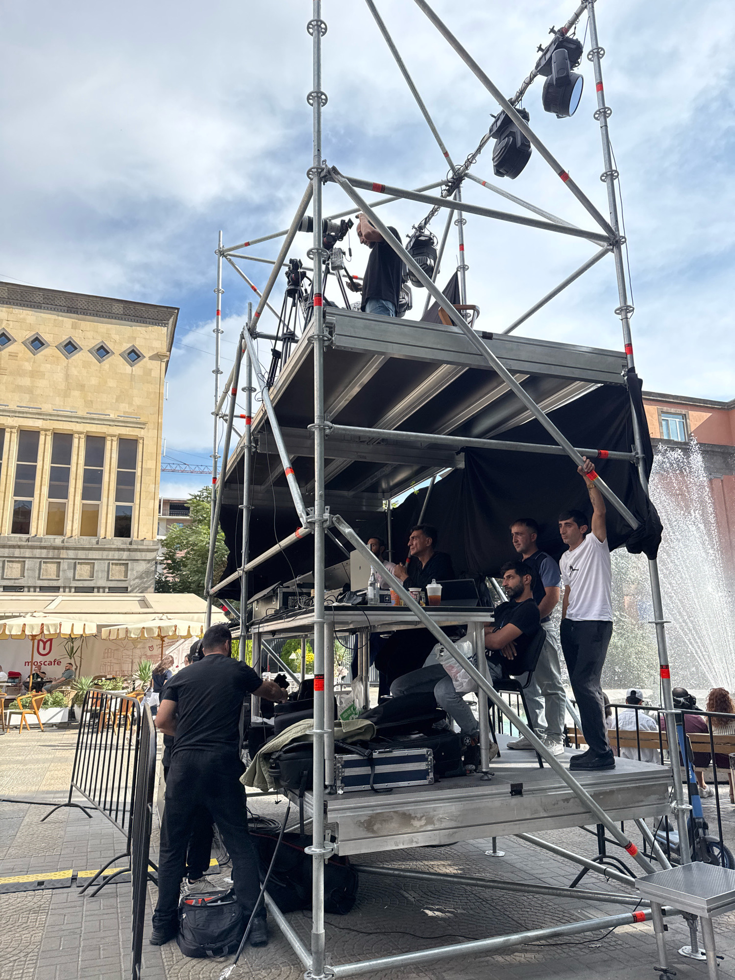 In Yerevan, Armenia, a temporary stage structure constructed of scaffolding and metal platforms is set up outdoors on a paved area. The structure hosts a media setup, including lighting equipment mounted on the upper levels and a control area on a lower platform. Several individuals are positioned on and around the structure, some appearing to be working with the equipment, others observing. In the background, a light-colored building with distinctive diamond-shaped windows and tall vertical windows is visible, along with some trees and a fountain. The scene suggests an event or performance is being prepared or taking place in a public urban space.