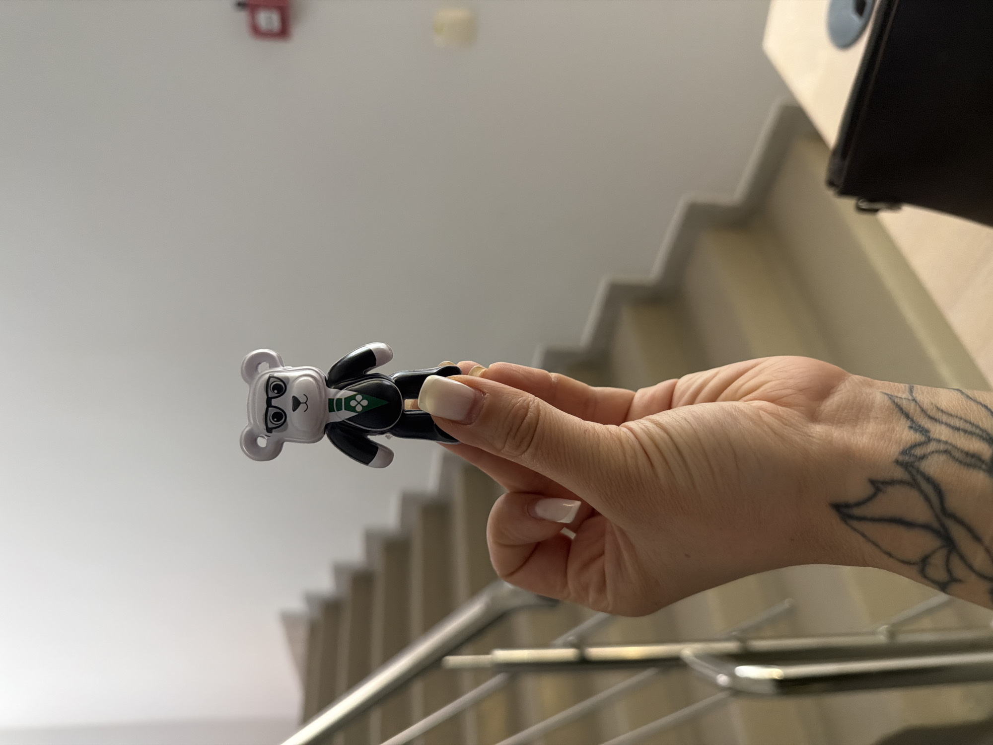 A person is holding a toy bear figure in front of a staircase. The toy bear is gray and black, wearing glasses and what appears to be a suit. The person's hand is holding the toy, and there is a tattoo on their wrist. The background shows the stairs and a white ceiling with what looks like a smoke detector. The location of Sarapul, Russia is not verifiable from the image.