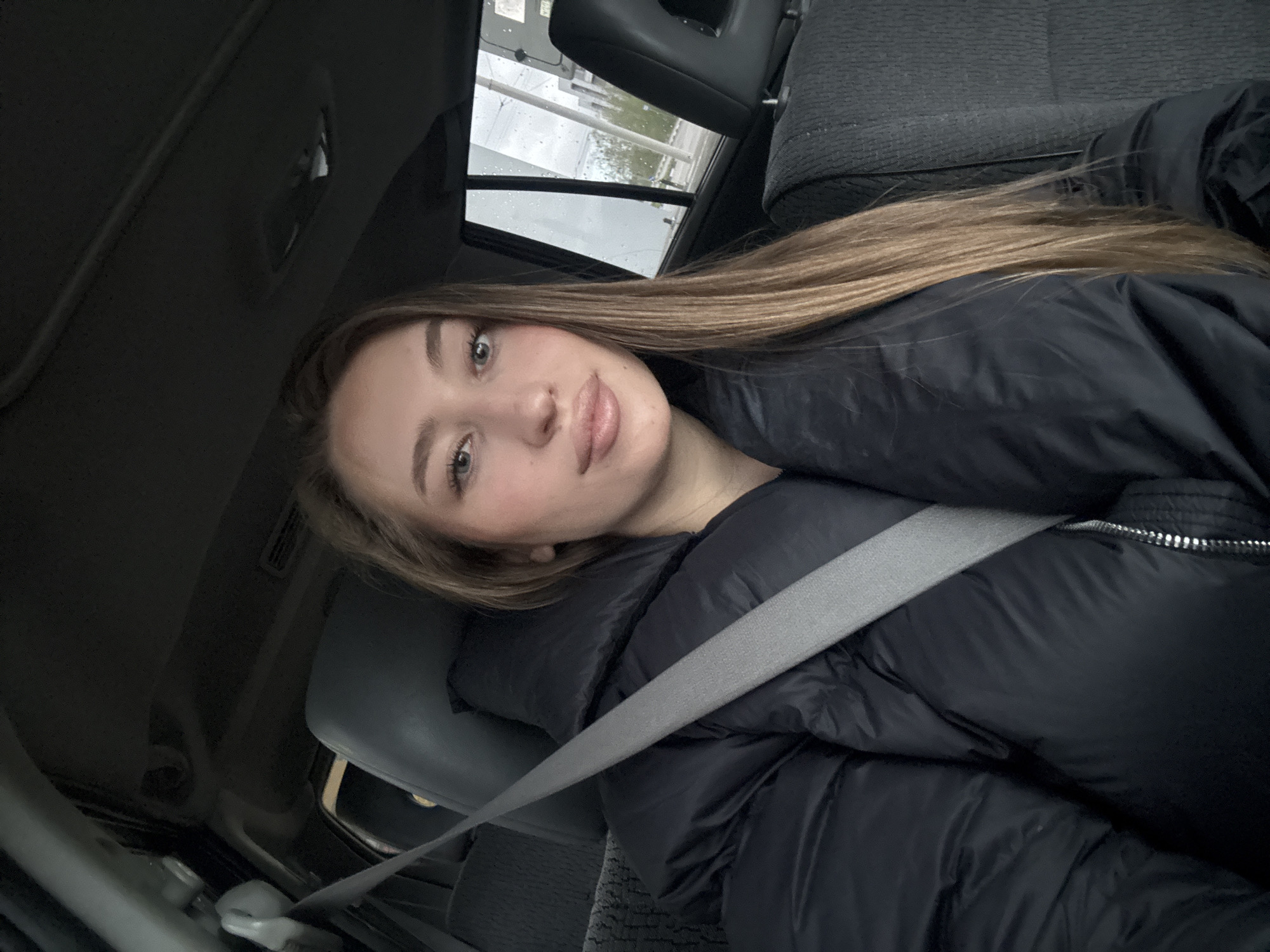 The image is a tilted, eye-level selfie of a young woman seated inside a car. She has long, straight, light brown hair that cascades over her right shoulder and across her back. Her light-colored eyes are directed towards the camera, and she displays a neutral expression with a slight, gentle smile. She is wearing a dark, possibly black, puffer jacket, and a gray seatbelt is fastened across her chest and left shoulder.
The car's interior features dark-colored seats, a headrest, and parts of the ceiling. Through the window behind her, an outdoor scene is visible. The lighting suggests it is daytime, but the sky is overcast, and the diffused light, possibly with raindrops on the window, indicates cloudy or rainy weather. Faint vertical structures are discernible outside, suggesting an urban or suburban environment, potentially a building or a gas station.
There is no visible text in the image. The scene captures a casual moment of the woman in a vehicle, located in Kharkiv, Ukraine, with a relaxed demeanor.