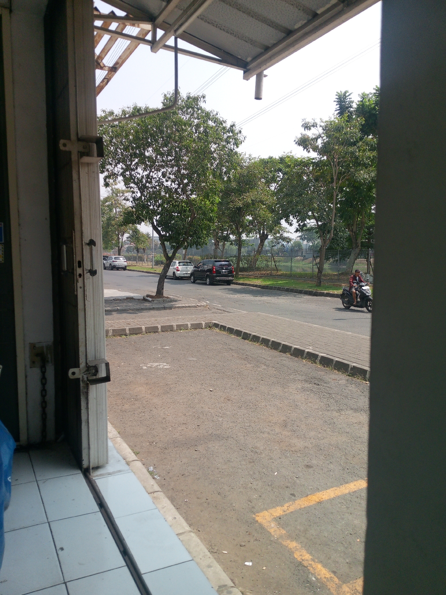 The image depicts a street scene in Cikupa, Indonesia, viewed from a doorway of a building. In the foreground, to the left, is the interior of a building with white tiled flooring and a partially visible blue object. A white metal door frame with a latch mechanism and a hanging chain is also prominent on the left. The doorway opens onto an outdoor paved area with a concrete curb.
Beyond the paved area, a road is visible with several vehicles. On the road, a black SUV is parked or driving, followed by a white sedan. Further down the road, a silver SUV is visible. A person is riding a scooter on the right side of the road, wearing a red jacket and dark pants.
The environment is characterized by a clear sky and lush green trees lining the roadside. A metal fence runs along the edge of a grassy area behind the road. Power lines are visible overhead. The overall impression is that of a typical urban or semi-urban street in Indonesia on a bright, sunny day.