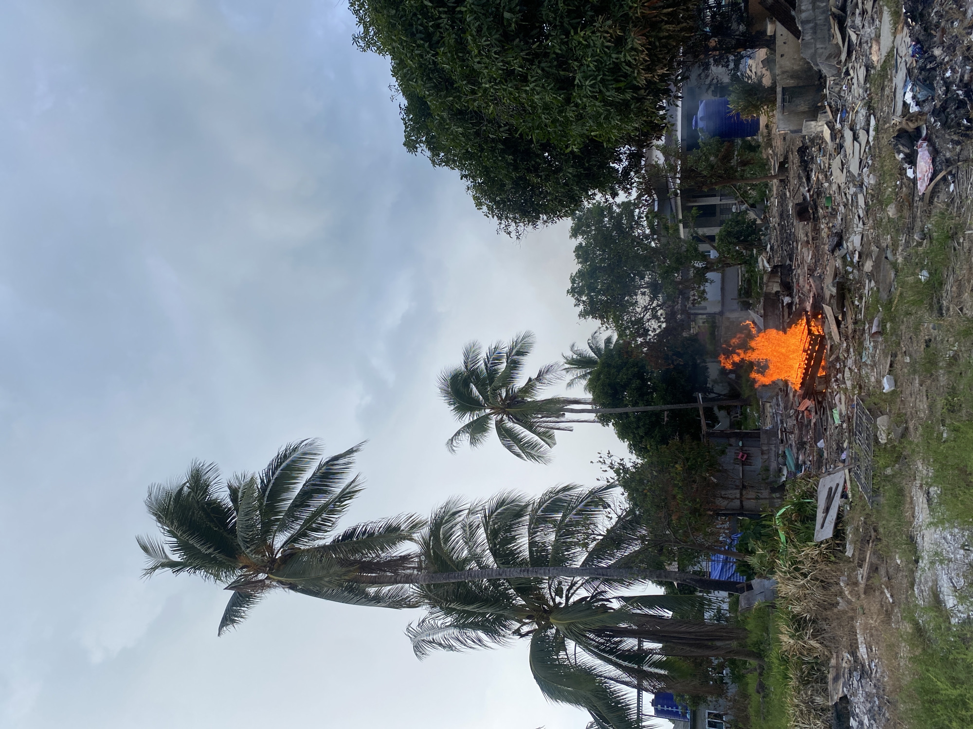 The image shows a scene in Baan Mae Nam, Thailand, with palm trees visible against an overcast sky. A fire is burning amidst debris and trash near a building and trees. The surrounding area appears to be littered and possibly damaged.