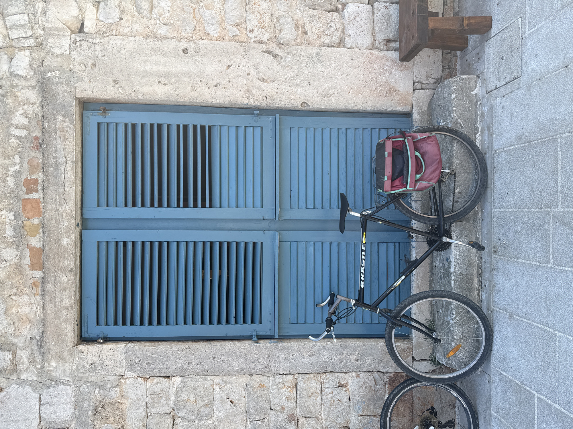 A bicycle with a red bag attached to the rear rack leans against a weathered stone wall in front of blue louvered shutters. The bicycle is black with silver handlebars and appears to be a mountain bike. Part of another bicycle's wheel is visible to the left of the main bicycle. The stone wall is constructed from rough-hewn blocks and shows signs of age and weathering. The shutters are a vibrant blue and are slightly ajar. A small, rustic wooden stool or bench is positioned to the right of the bicycle. The ground in front of the scene is paved with grey stone tiles. The overall impression is of a rustic, possibly Mediterranean, setting, consistent with a location like Jelsa, Croatia, known for its stone architecture and charming streets.