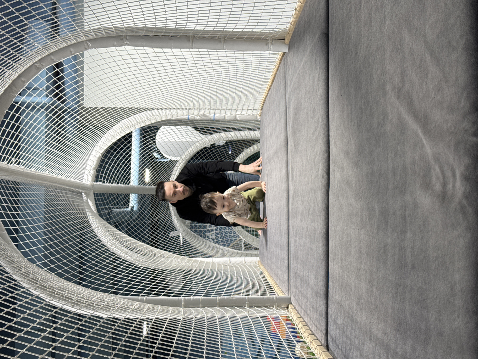 A low-angle shot reveals a man and a young child situated within a play structure, possibly indoors, in the city of Izhevsk, Russia. The child, wearing a light-colored patterned shirt and darker pants, is positioned closer to the right side of the frame, his hand grasping the edge of a padded, light-gray surface. The man, appearing to be the child's father, is slightly behind and to the left, dressed in a black long-sleeved shirt and blue jeans. The play structure is defined by interconnected white netting creating a tunnel-like space. Background lighting hints at the room's brightness.