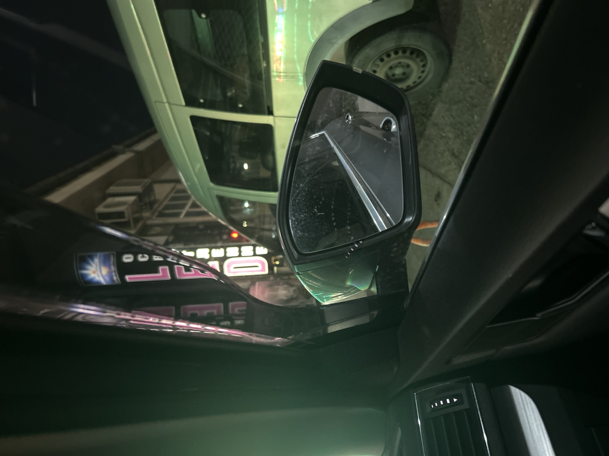 The image is taken from inside a car, focusing on the rearview mirror. Reflected in the mirror is a light-colored van with its tire visible. Outside the car, there is a building, and a sign with the word "DEAD" is partially visible. The image provides no additional evidence that it was taken in Burgas, Bulgaria.