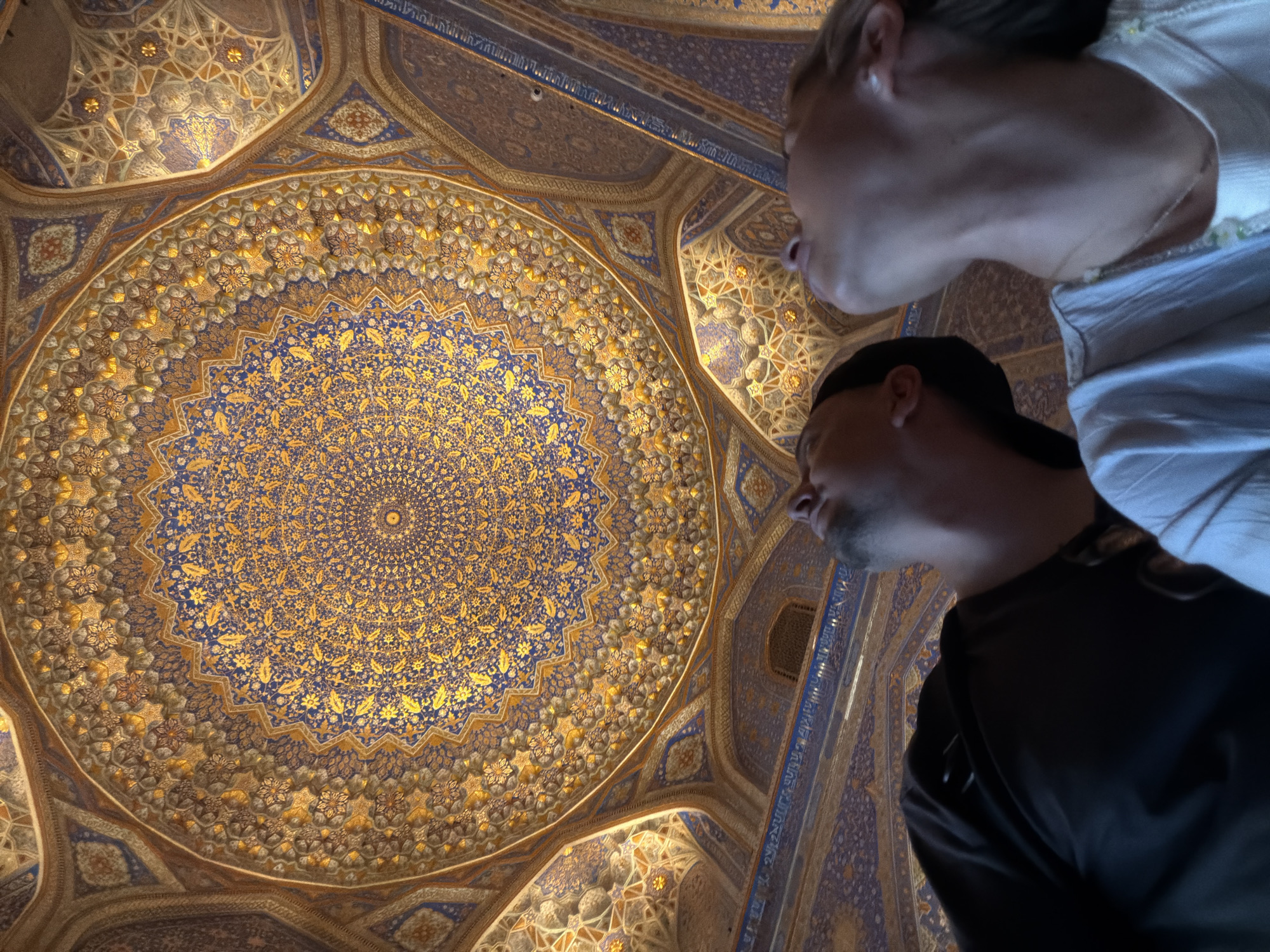 The image depicts the interior of a grand building with an intricately decorated dome in Samarkand, Uzbekistan. The dome is adorned with a complex geometric pattern featuring shades of blue and gold, with smaller floral motifs. The walls and other ceiling structures also display elaborate blue and gold tilework. In the foreground, two individuals are visible from the chest up, looking upwards towards the dome. The person on the left is wearing a black shirt, and the person on the right is wearing a light blue or white shirt with a necklace. The perspective is from below, emphasizing the scale and grandeur of the architecture.
