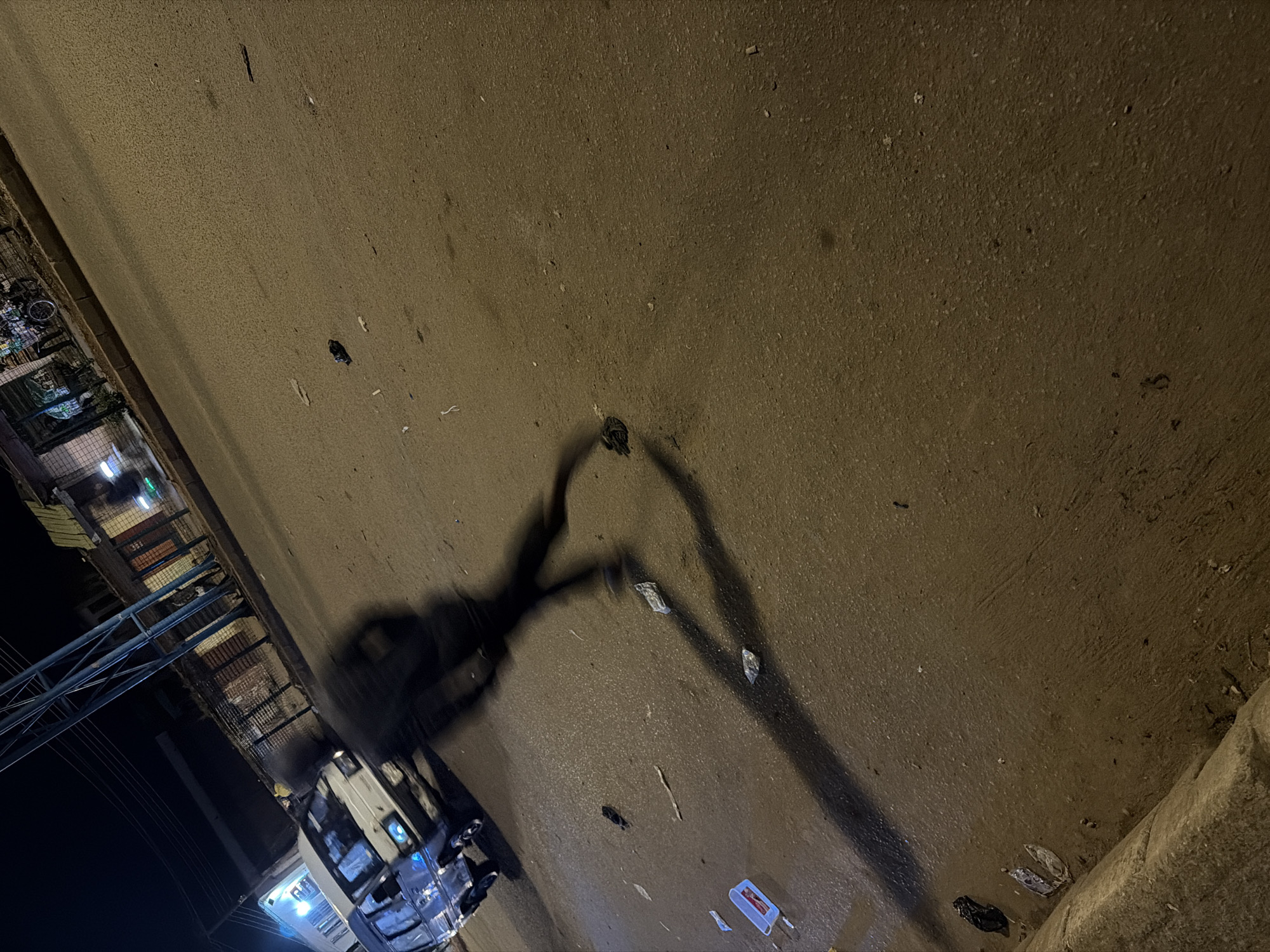 In Bukuru, Nigeria, at night, a blurred figure in dark clothing is captured in motion on a paved road. The figure, appearing to be running or walking briskly, casts a long shadow on the asphalt. Scattered across the road are various pieces of debris, including what appear to be crumpled papers and plastic containers. To the left of the frame, a white van is parked. In the background, a dimly lit street with buildings and a metal structure are visible. The overall scene suggests a dynamic moment on a gritty, urban street at night, with the motion blur of the person being the central focus against the static elements of the environment.