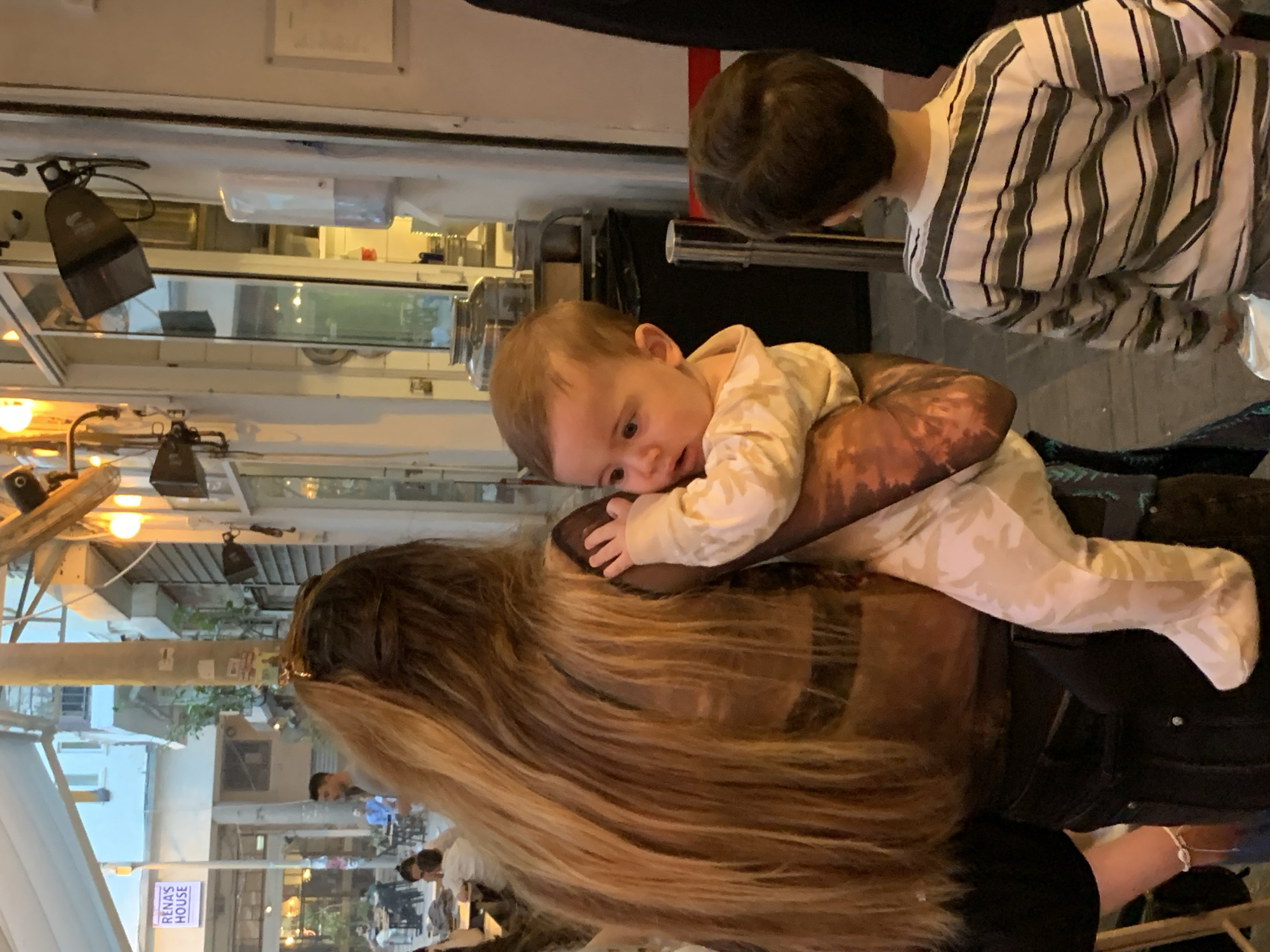 A woman with long blonde hair holds a baby in her arms. Another child stands nearby wearing a striped shirt. The scene appears to be outdoors at night in Tel Aviv, Israel, as evidenced by a sign in the background that reads "Rena's House". Other people are sitting at tables nearby. The lighting is a combination of natural and artificial light sources.