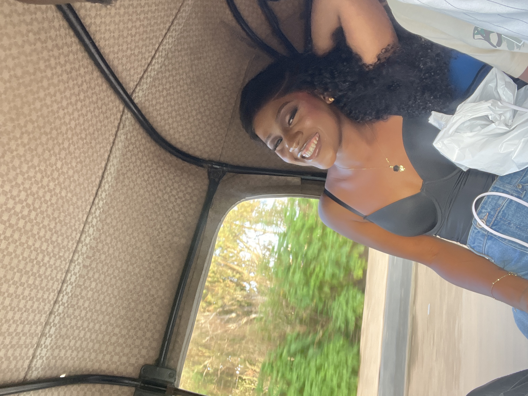 A woman with dark skin and long, curly black hair smiles broadly, looking towards the camera from the interior of a moving motorized tricycle, likely a Keke Napep, in Gura Top, Nigeria. She is wearing a black spaghetti strap top, blue denim jeans, a gold necklace with a black flower-like charm, gold stud earrings, and a thin gold bracelet on her left wrist. Her right arm is positioned behind her head. The vehicle's ceiling is light beige with a subtle checkerboard or woven pattern, and black metal structural bars are visible. Through the open window on the left, a blurred background of green foliage, trees, and bright sky indicates motion. A white plastic shopping bag is visible in the lower right foreground, and a partial view of another person's arm wearing a light-colored, vertically striped shirt is on the far right.