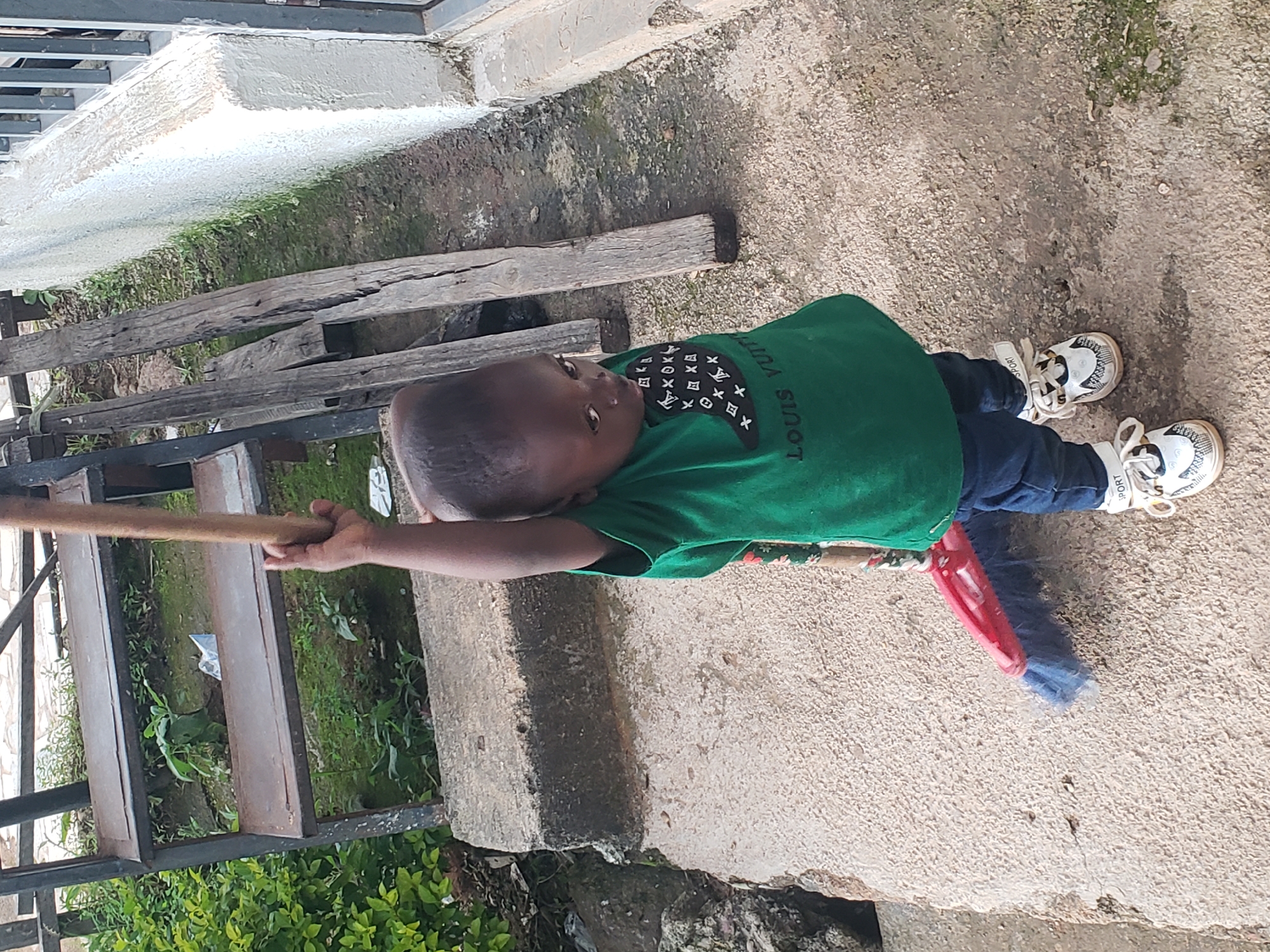 In Jos, Nigeria, a young child is captured in a full-body shot, standing on a concrete surface and holding a broom upright. The child, wearing a green t-shirt and blue jeans, has a playful expression as they look towards the camera. The broom, with a red handle and blue bristles, is positioned behind the child, suggesting a moment of rest from cleaning. The background reveals a slightly overgrown area with plants and some discarded wooden planks, indicating a residential or semi-developed outdoor setting in the city.
