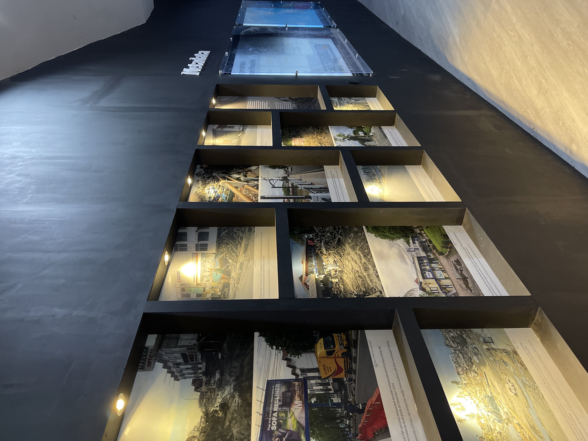 This image depicts an exhibition on the wall of a dark room, likely within a museum or cultural center in Banda Aceh, Indonesia. The exhibition features a series of framed photographs displayed within recessed shelves and on the wall itself. The photographs appear to document the aftermath of a disaster, likely the 2004 Indian Ocean tsunami, given the location.
In the foreground, the wall is painted black and illuminated by spotlights, casting shadows on the floor. The floor is a light-colored wood-look laminate. The exhibition is arranged in a grid-like fashion.
On the left side of the wall, several photographs show scenes of destruction: collapsed buildings, debris-strewn streets, and damaged infrastructure. One photograph prominently features a billboard for "SOFA RECLINER" with a brand name "Mmedia". Another shows a yellow delivery truck on a street.
Further to the right, more photographs continue to display the devastation. There are images of buildings that are partially submerged or severely damaged, with debris piled high. The photographs are accompanied by captions, though they are too small to read in detail.
On the right side of the wall, there are larger, framed acrylic panels, also displaying text and images, but these are less clear from this perspective. Above the exhibition, a raised, illuminated word "Metadata" is visible on the black wall.
The overall impression is one of remembrance and documentation of a significant historical event in Banda Aceh.