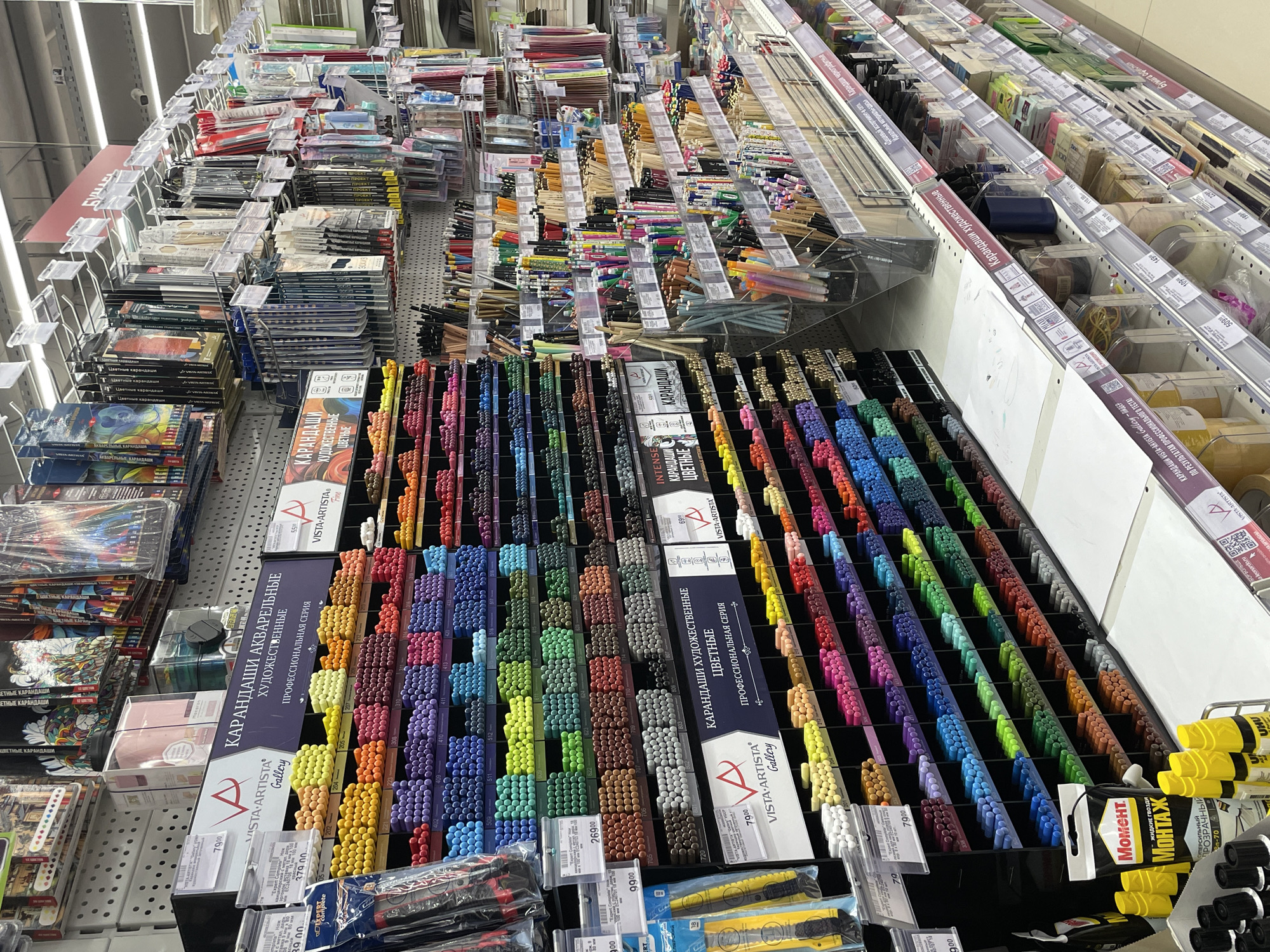 The image shows a well-stocked art supply section in what appears to be a retail store in Moscow, Russia. Shelves are filled with various art materials, including watercolor pencils and colored pencils from the brand "Vista Artista Gallery". The pencils are arranged by color and have price tags displayed. Other items like glue and paper are also visible.