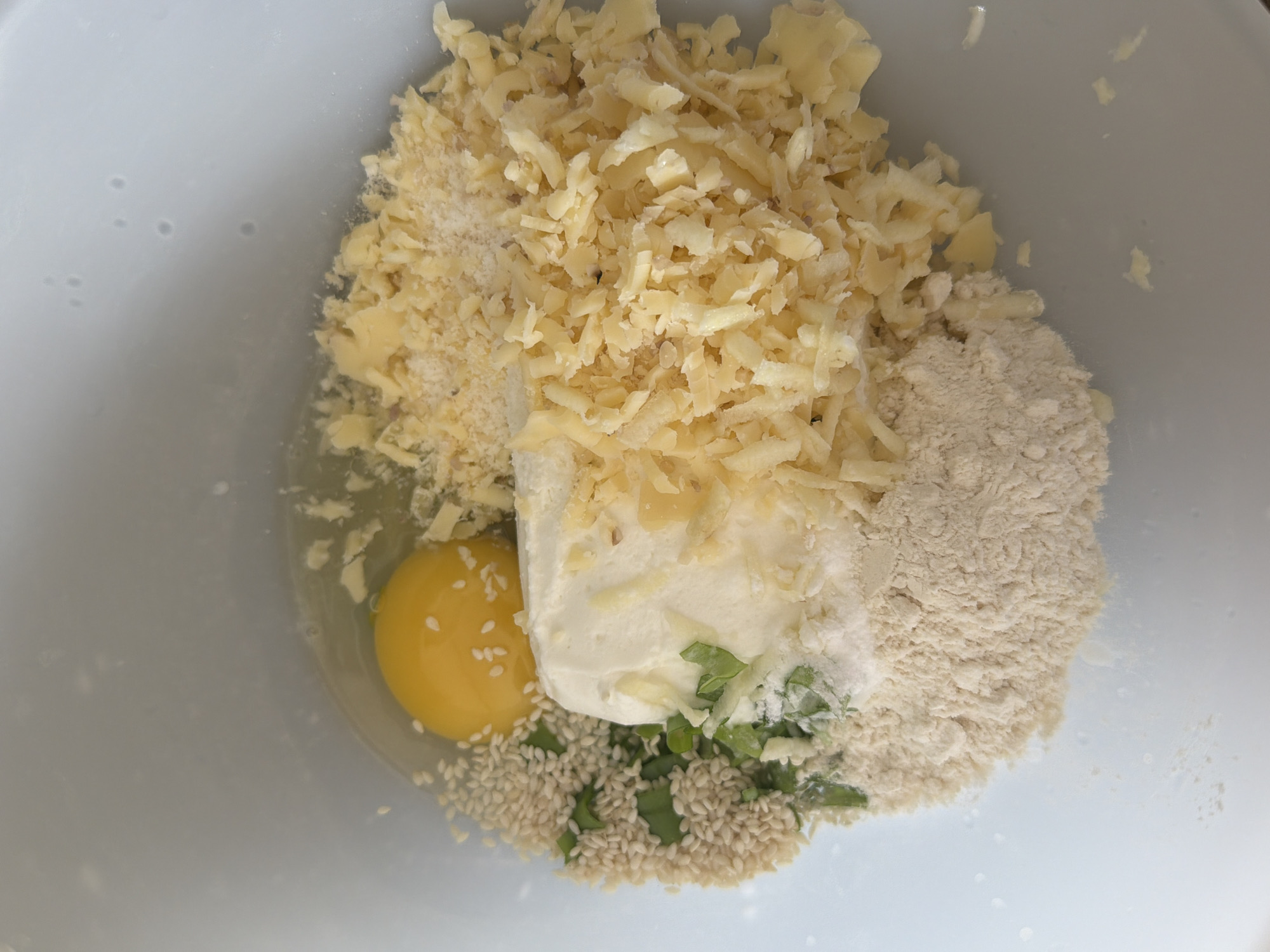 The image displays a close-up, top-down view of various food ingredients gathered inside a white mixing bowl, awaiting preparation. The primary subject is a collection of unmixed ingredients.
The most prominent ingredient is a large pile of light yellow, irregularly shredded cheese, possibly a hard or semi-hard variety, occupying a significant portion of the bowl. Mixed within and alongside this shredded cheese are smaller sprinkles of a finer, white grated substance, likely Parmesan cheese or similar.
To the bottom left of the cheese, a single raw egg with an intact bright yellow yolk and clear albumen is visible. Adjacent to the egg and below the main cheese pile is a generous spoonful or dollop of a creamy white substance, which appears to be a soft cheese like cream cheese or ricotta.
Below the creamy substance and to the left, a heap of small, pale, oval-shaped white sesame seeds is spread out. Interspersed among the sesame seeds and partially covered by the creamy cheese are finely chopped green herbs, resembling chives or parsley.
To the right side of the creamy cheese and extending towards the top right, there is a pile of light brown or beige powdery substance, consistent with flour, almond flour, or breadcrumbs.
The ingredients are distinctly separate, indicating they are in the initial stages of being combined for a recipe. The overall scene suggests food preparation in a domestic kitchen setting. No people, visible text, specific time of day, or weather conditions are discernible from the image. The location context provided is Tel-Aviv, Israel.