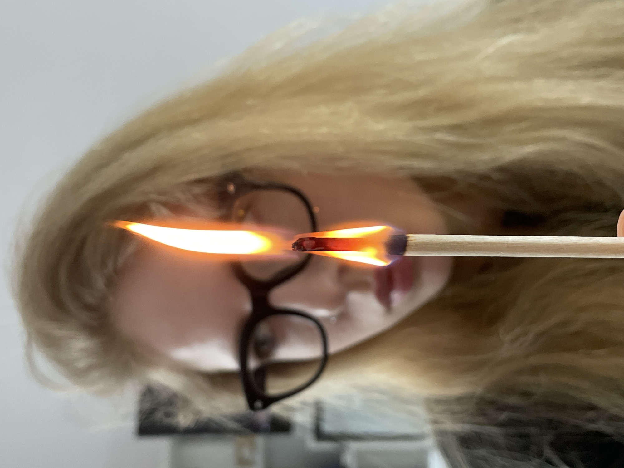 A young woman with blonde hair and glasses holds a lit match in front of her face. The flame is prominent, obscuring some of her features. The background is a blurred, neutral tone with some indistinct objects. The location is not identifiable as Izhevsk, Russia based on the image content.