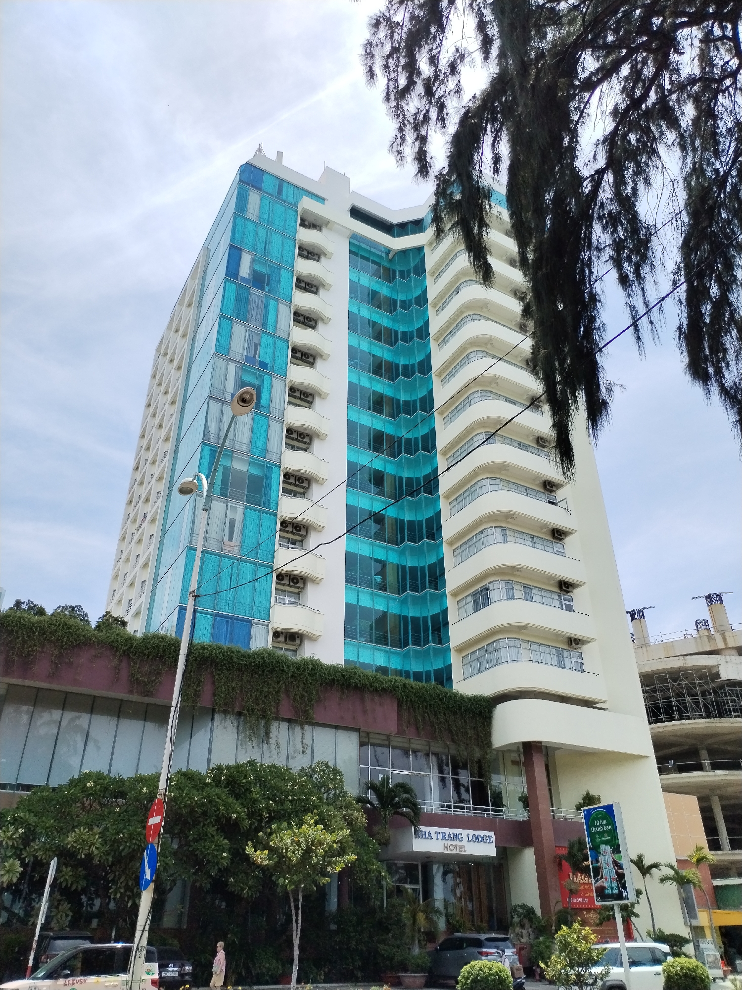 This is a medium shot of the Nha Trang Lodge Hotel in Nha Trang Ward, Vietnam. The hotel is a tall, multi-story building with a modern design. The left side of the building is covered in blue glass windows, while the right side has white balconies and windows. In the foreground, there are trees and a street with a few cars. A street lamp with two lights stands in the middle of the frame. A woman in a pink shirt and beige shorts is walking on the sidewalk. There are construction cranes in the background, indicating ongoing development in the area. The sky is cloudy.