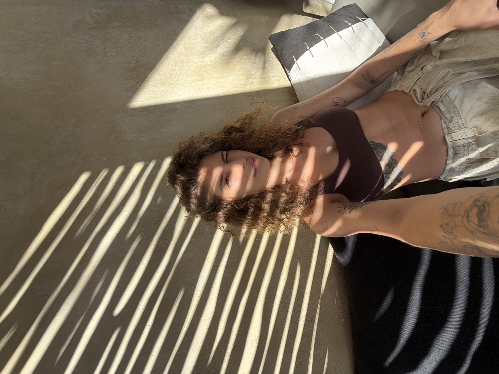A person with curly brown hair is captured in a selfie taken outdoors in a sunlit environment. The individual is wearing a dark brown bikini top and light-colored, loose-fitting pants. Tattoos are visible on their arm and torso. The background features a textured, light-colored wall with strong diagonal shadows cast by what appears to be blinds or foliage. A dark sofa is partially visible in the lower left of the frame, and a pillow with a black and white geometric pattern rests on a couch to the right. The scene suggests a relaxed, possibly tropical setting, possibly in a cafe or resort area. It is difficult to definitively ascertain the location as Mengwi, Indonesia, from the visual information alone; however, the style of the architecture and the lighting are consistent with outdoor spaces found in tropical regions.