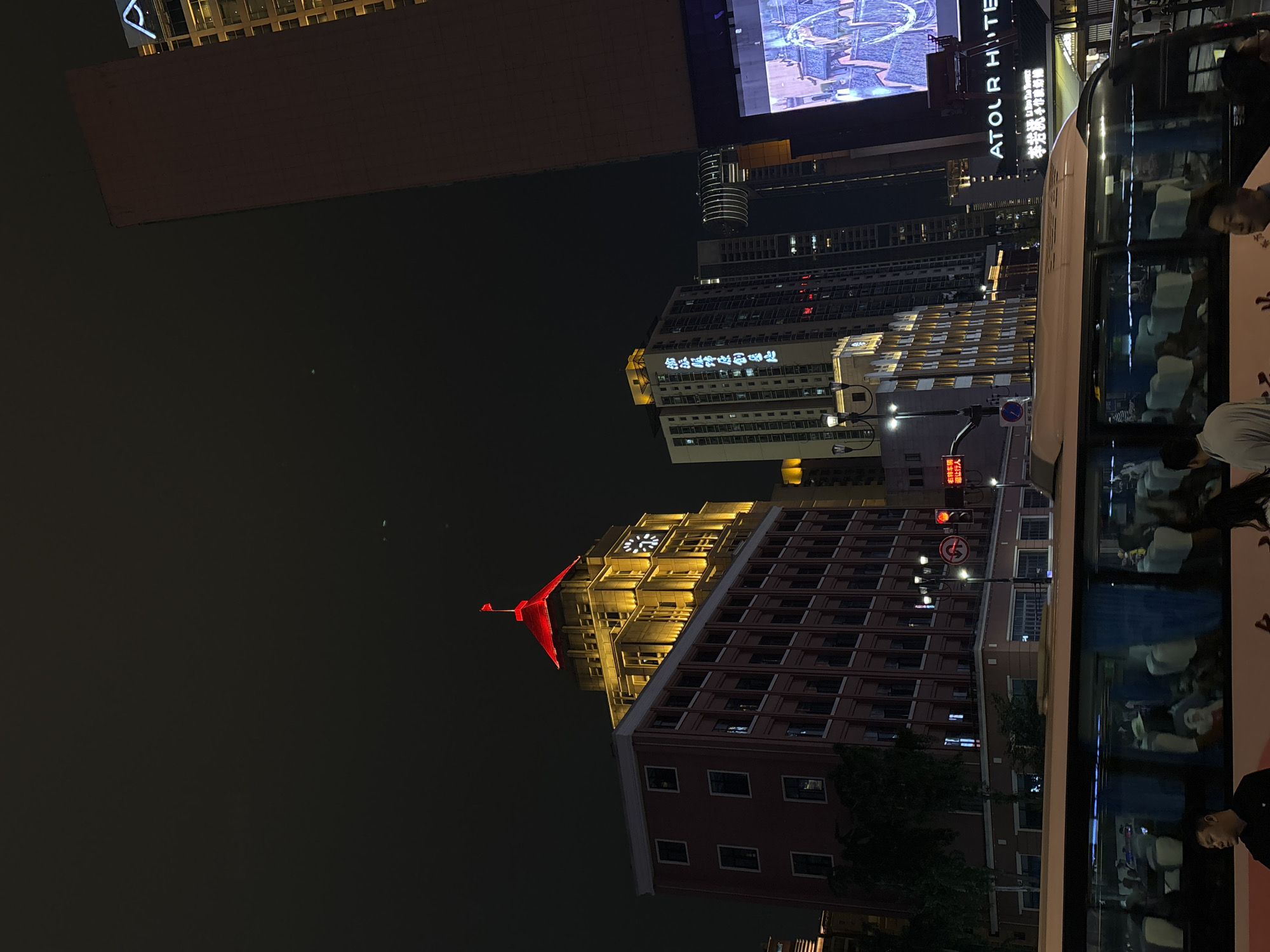 This media file captures a vibrant urban street scene at night in Yuzhong District, China. The setting is illuminated by numerous artificial lights from buildings, signs, and street infrastructure, contrasting sharply with the dark night sky.
Several tall buildings dominate the cityscape. On the right side of the image, a large white bus is visible, its windows reflecting the surrounding lights and showing faint outlines of passengers inside. Just behind the bus, a prominent building features a brightly lit sign for "ATOUR HOTEL" (亚朵酒店). Above this, a large digital billboard displays intricate patterns, possibly an advertisement.
Further into the background, other buildings are illuminated, including one with bright Chinese characters that appear to read "重庆日报" (Chongqing Daily), suggesting a media or newspaper office. To the left, a distinct, older-style building stands out, topped with a striking red, abstract, bird-like sculpture and an illuminated clock face. This building's upper facade is brightly lit with warm yellow light.
A dark body of water, likely a river or large canal, occupies the far left portion of the image, reflecting some of the distant city lights. On the street, a streetlight and various traffic signs are visible. One sign, red with white Chinese characters, states "右转待转区 请行人注意" (Right turn waiting area, pedestrians please pay attention). People are seen near and within the bus, indicating ongoing evening activity. The overall impression is one of a bustling, modern city alive at night.