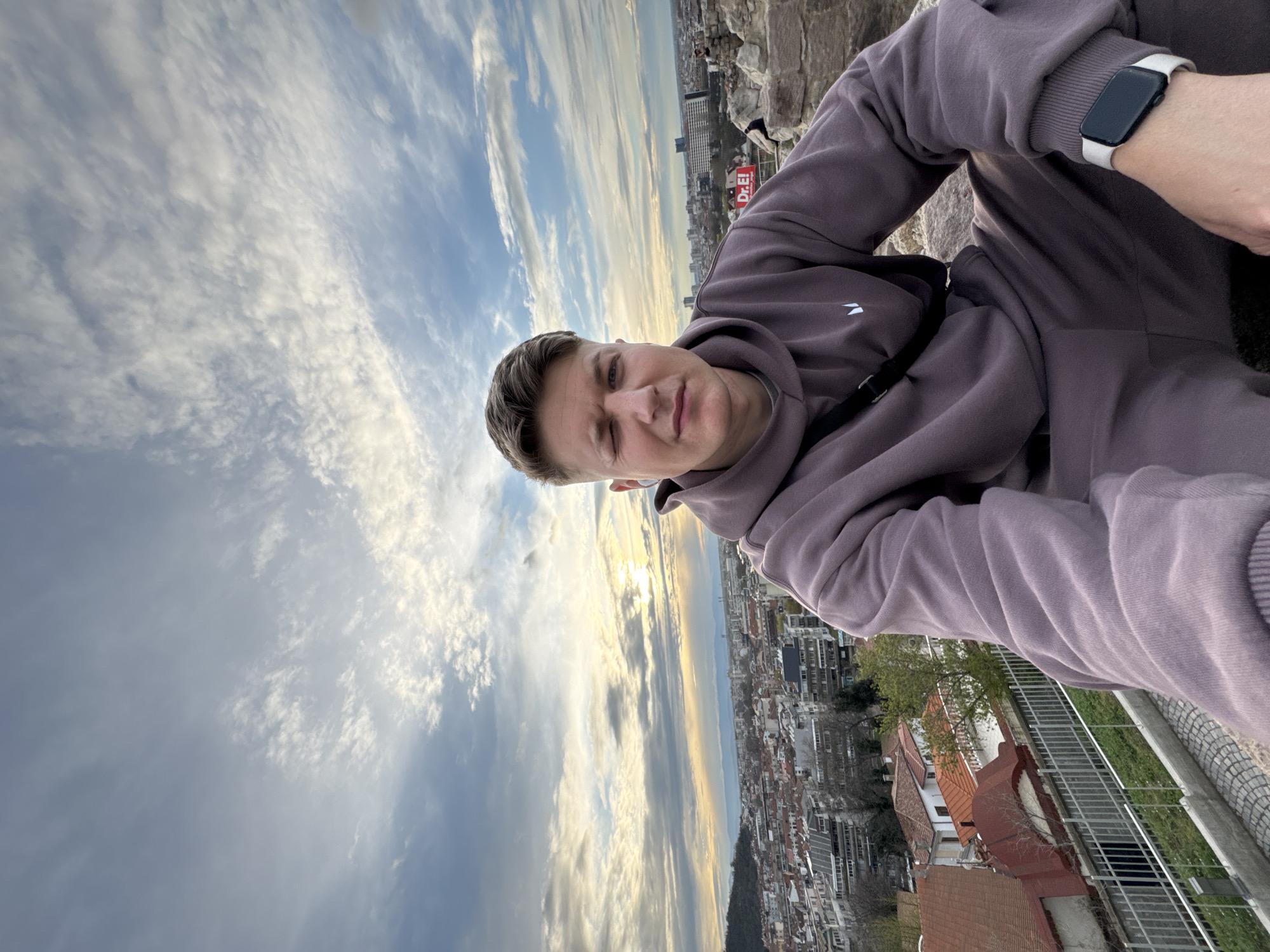 A man is shown from the chest up, wearing a mauve-colored hoodie with a white logo on the left breast and a white smartwatch on his left wrist. He has short, light brown hair and is looking into the camera with a slight smile. Behind him is a cityscape with numerous buildings and rooftops. The sky is partly cloudy with a mix of blue and white, and the sun is visible near the horizon. The photograph is rotated counter-clockwise and was likely taken at the top of Bunardzhik Hill, also known as the Liberators Hill, in Plovdiv, Bulgaria.