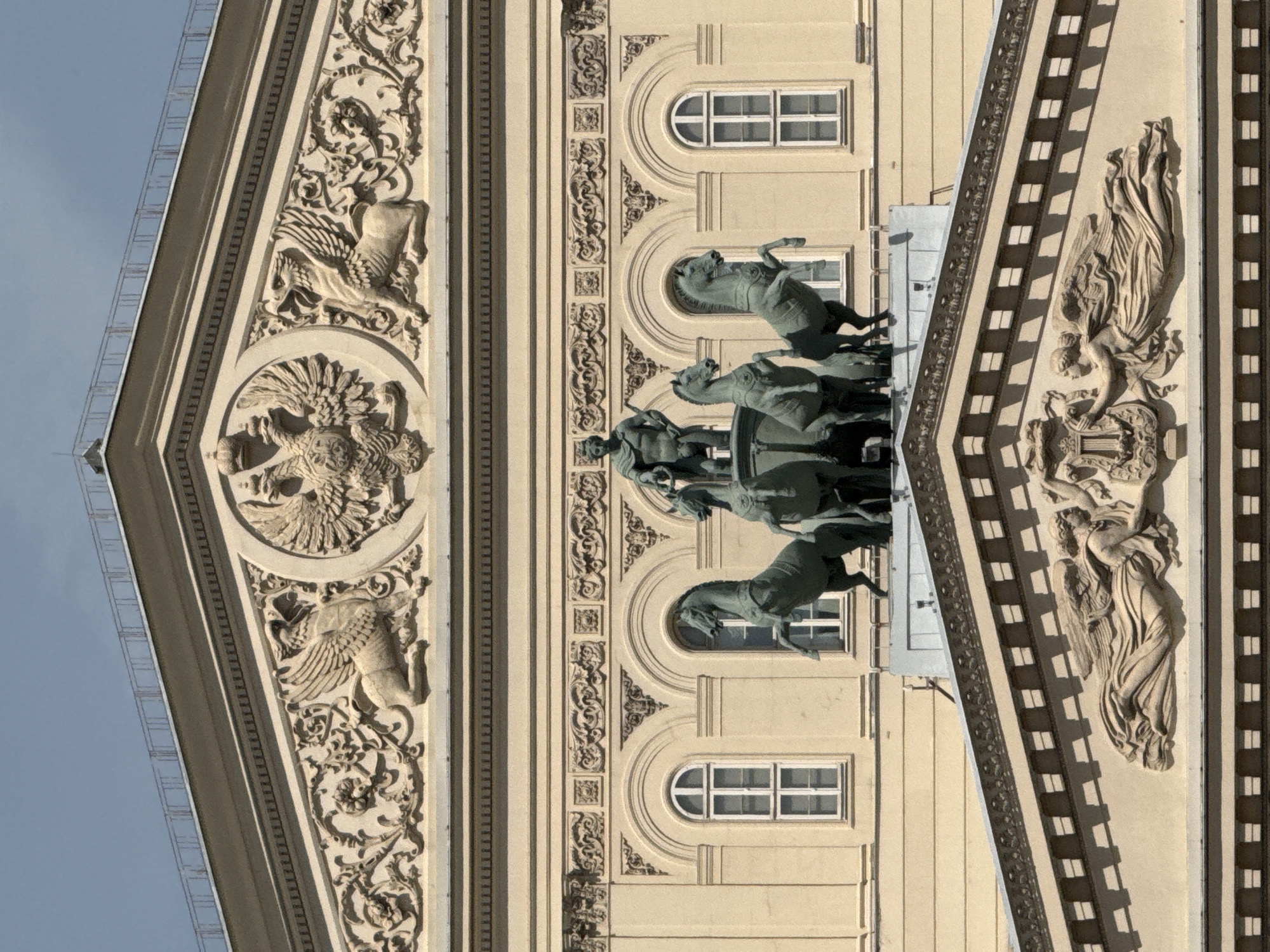 The image depicts architectural details of the Bolshoi Theatre in Moscow, Russia. The facade is adorned with ornate carvings, including winged creatures, floral patterns, and classical figures. A prominent sculpture of a chariot pulled by horses stands out. Windows with arched tops are visible in the building's background. The sky is pale blue.