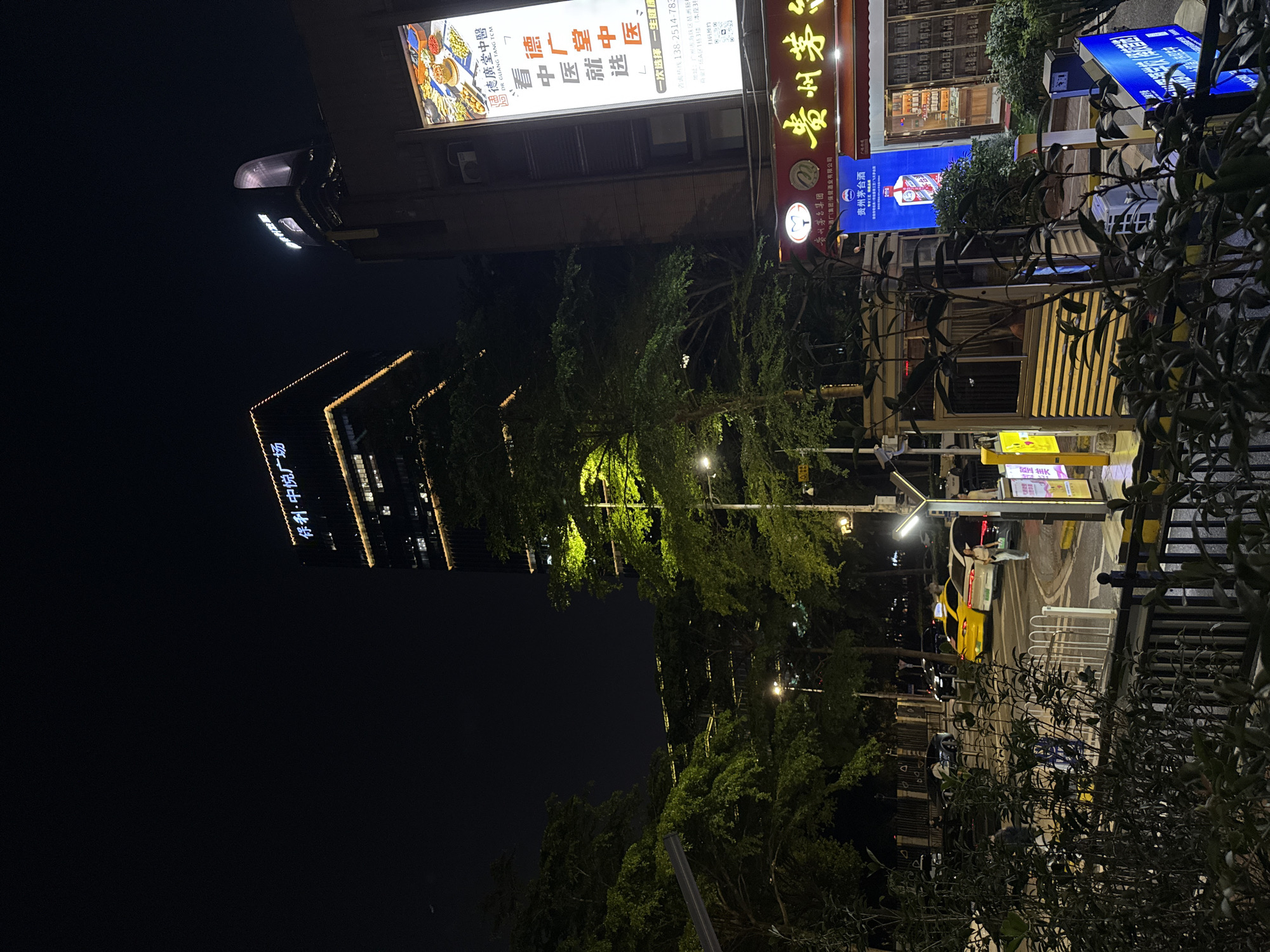 The image captures a vibrant night scene in an urban area, depicting a street lined with buildings and illuminated by various signs and streetlights. The perspective is from street level, looking towards a tall skyscraper in the background and commercial establishments closer to the foreground.
**Subjects:**
The primary subjects are buildings of varying heights, including a prominent skyscraper. Numerous trees with lush green foliage are visible, some partially lit. A street with vehicles, including a distinct yellow taxi, and at least one pedestrian, occupies the middle ground. Streetlights, overhead wires, and fences complete the urban landscape.
**Activity or event taking place:**
The scene shows typical nighttime activity in a commercial district, with businesses open and lit up. People and vehicles are present, indicating a regular evening in the city.
**Notable details:**
The **time of day** is clearly night, as indicated by the dark sky and the reliance on artificial lighting from signs, streetlights, and building interiors. The **weather** appears clear.
**Location cues** are abundant through the Chinese text on the signs, confirming an East Asian setting. The provided context specifies the **location** as Haizhu District, China.
**Visible text** includes:
* On the tall skyscraper: "保利·中悦广场" (Poly China Joy Plaza).
* On a large, bright billboard: "德廣堂中醫" (De Guang Tang TCM - Traditional Chinese Medicine), accompanied by slogans such as "看中医就选德广堂中医" (Choose De Guang Tang TCM for Traditional Chinese Medicine) and "一次选择一生健康" (One choice, lifelong health). Contact information ("咨询热线138-2514-783") and an address in "广州市海珠区洲新商业广场A区13楼" (13th floor, A District, Zhousin Commercial Plaza, Haizhu District, Guangzhou City) are also present, along with "扫码预约" (Scan code to book).
* Another sign advertises "贵州茅台集团" (Guizhou Moutai Group) and "贵州茅台" (Guizhou Moutai), a famous Chinese liquor brand, noting "酒厂(集团)保健酒业有限公司" (Winery (Group) Health Wine Industry Co., Ltd.).
The image conveys the lively and well-lit atmosphere of a commercial area in a Chinese city after dark.