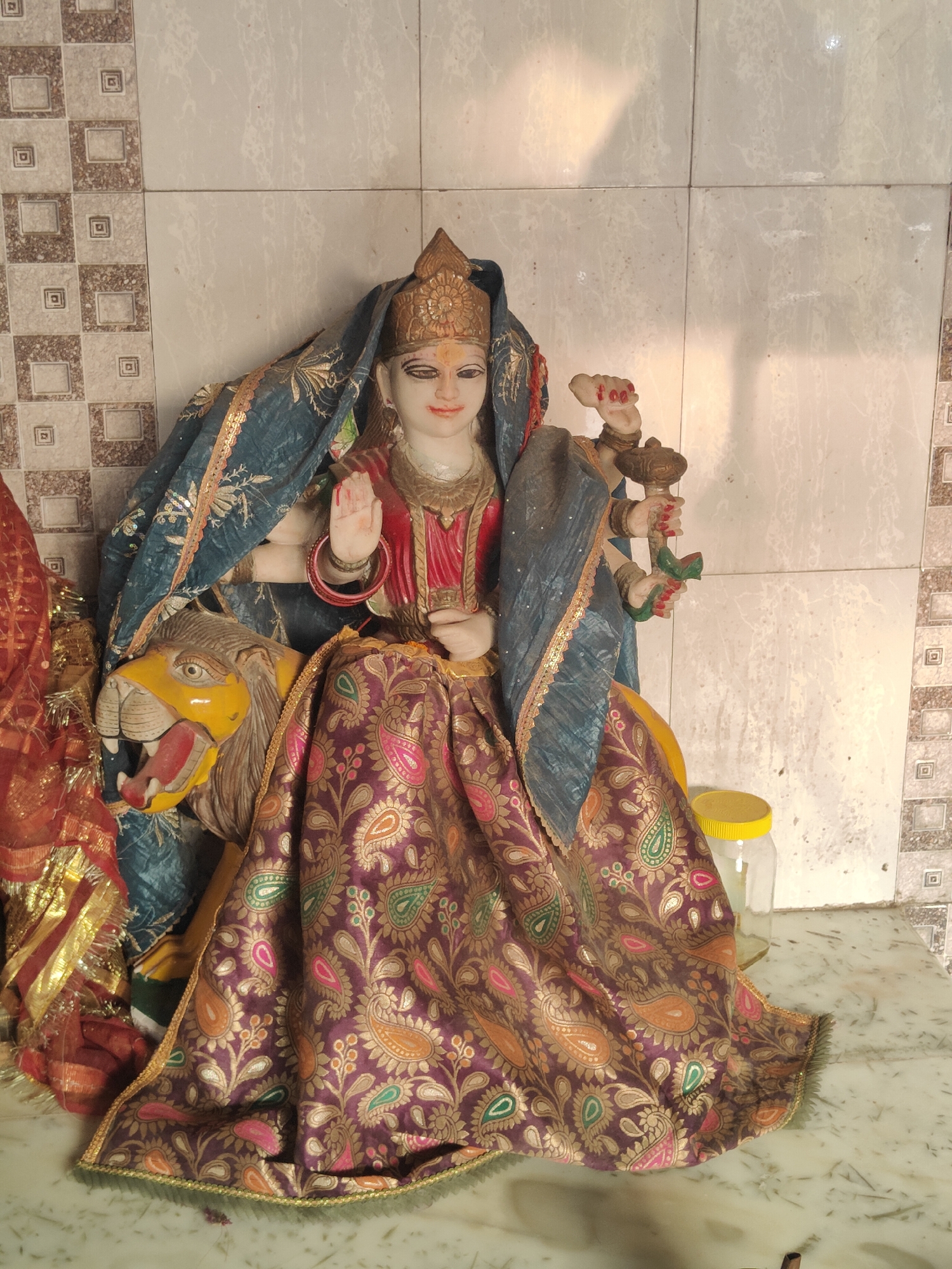 The image shows a statue of the Hindu goddess Durga seated on a lion, a common iconography for the deity. The statue is adorned with a blue and gold draped fabric and a richly patterned skirt. She holds several objects in her hands, suggesting different divine attributes. The backdrop is a tiled wall, with a decorative pattern on the left side. A plastic jar with a yellow lid is visible to the right of the statue. The statue is placed on a marble surface, which is common in temples and religious spaces. This scene is likely from a Hindu temple or shrine. Without specific identifying landmarks or contextual information within the image itself, it is impossible to definitively place this scene in Gurgaon, India. However, such religious imagery is prevalent throughout India, including Gurgaon.