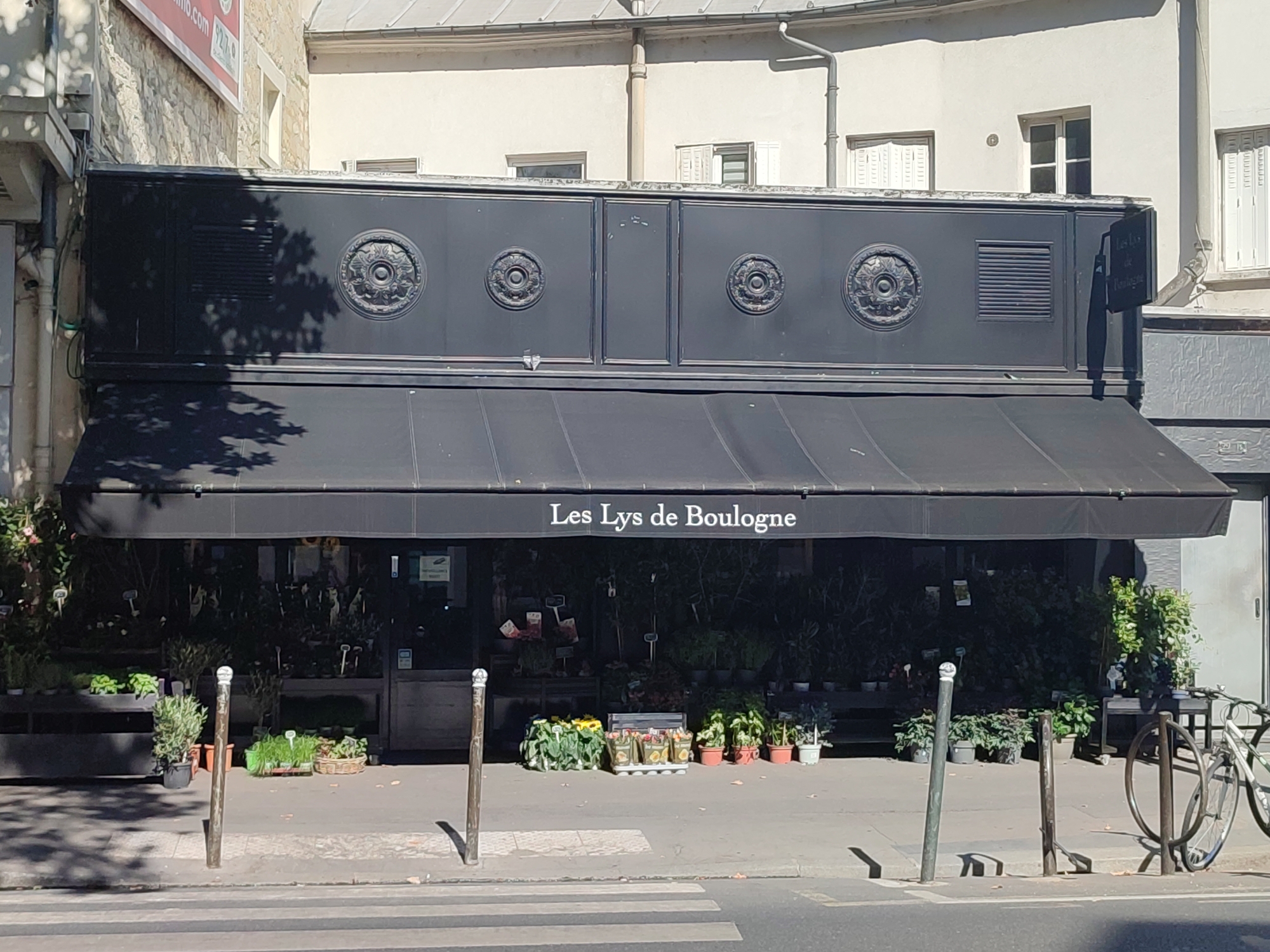 The image shows a storefront in Boulogne-Billancourt, France, with the name "Les Lys de Boulogne" displayed on the awning. The shop appears to be a flower or plant shop, with numerous plants and flowers displayed outside on the sidewalk. The storefront itself is dark-colored with decorative circular elements. The street in front of the shop has a crosswalk, and a bicycle is parked to the right. The surrounding buildings are light-colored with windows and shutters. The scene is bathed in sunlight, casting shadows from trees and objects onto the sidewalk.