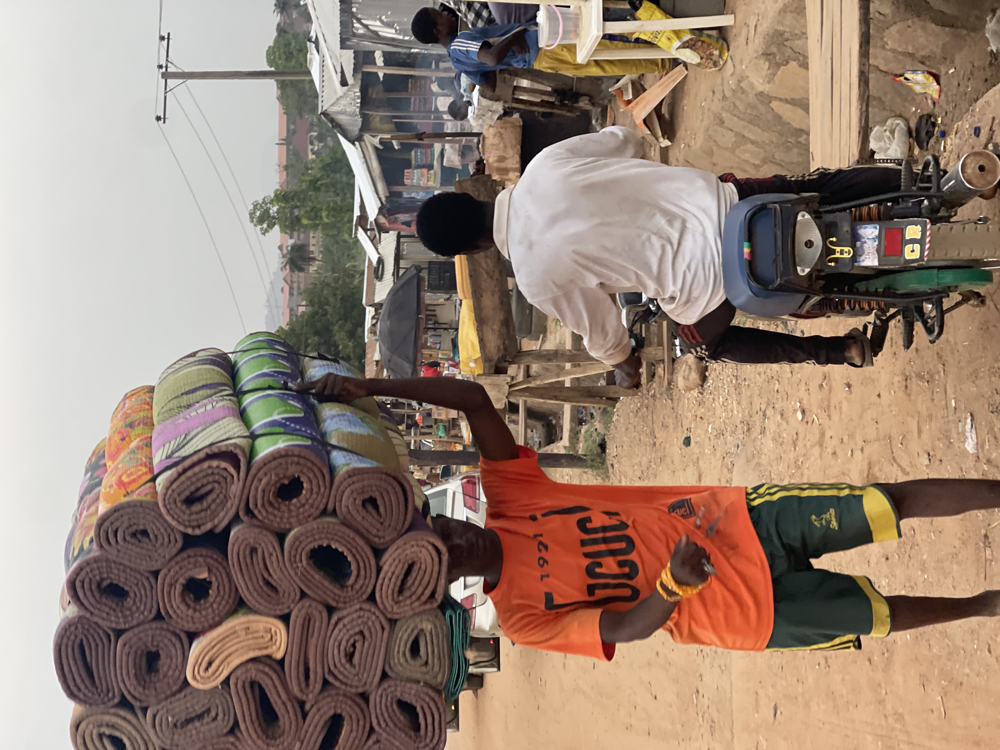 A medium-shot photograph of a man carrying rolled mats on his head in what appears to be a market environment in Abuja, Nigeria. The man is wearing an orange shirt with "OCUC" and "1921" printed on it, and green shorts with yellow stripes. He is carrying a stack of rolled mats in various colors and patterns on his head. His arm is raised, possibly to stabilize the load. The background shows other people, market stalls, a motorcycle, and a dirt road, suggesting an active commercial area. Buildings and utility poles are visible in the background.