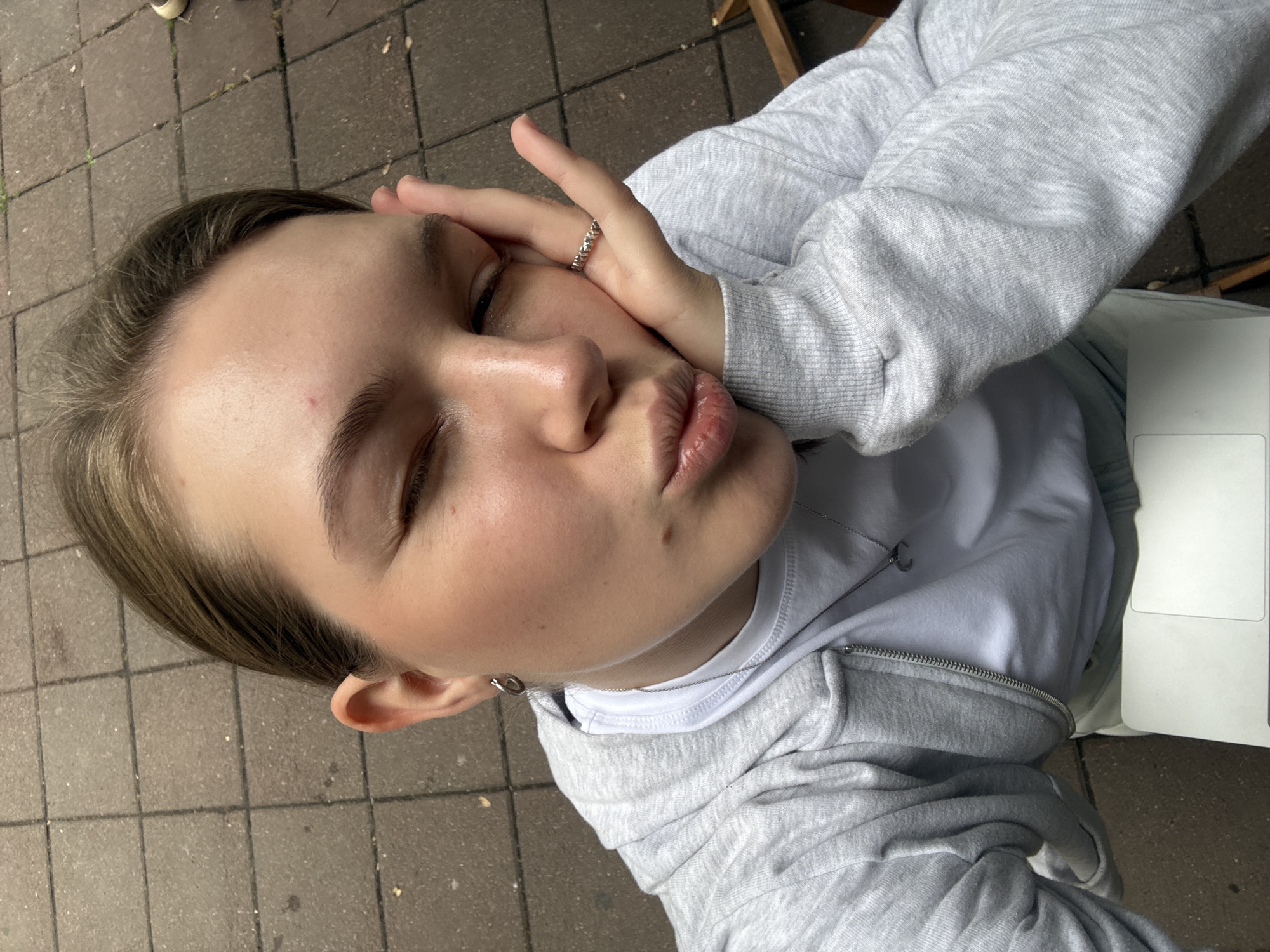 A young woman, with blonde hair pulled back, is captured in a close-up selfie. She is wearing a grey hooded sweatshirt over a white t-shirt and a silver necklace with a crescent moon pendant. A silver ring with small charms adorns her right hand, which is propping up her cheek. Her left eye is partially closed, and her lips are puckered. The background suggests an outdoor urban setting with a paved ground composed of square tiles. A portion of a laptop is visible in the lower part of the frame. The lighting is natural, suggesting daytime. The scene does not offer any specific clues to identify the location as Belgrade, Serbia.
