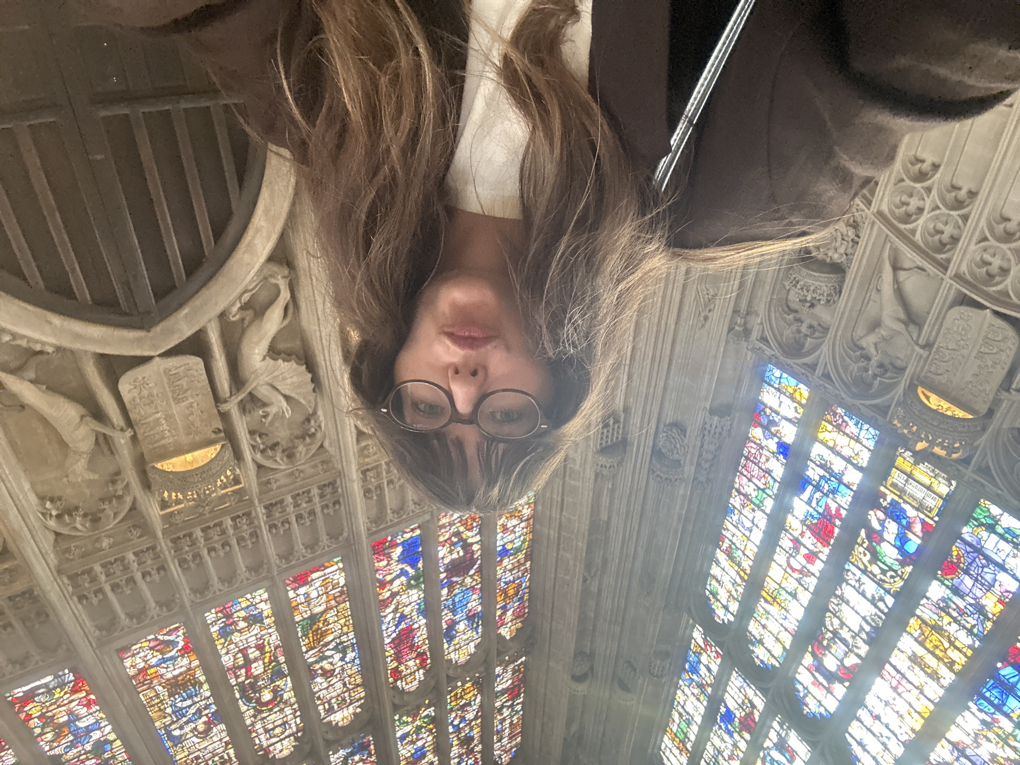 A low-angle selfie is taken by a young woman with long, brown, wavy hair and glasses. She is wearing a dark brown jacket over a white top and is looking towards the camera with her lips slightly pursed. Behind her, a grand setting features a tall, ornate stained-glass window with vibrant colors and intricate patterns, characteristic of Gothic architecture. The walls are made of stone and are adorned with carved reliefs, including what appears to be a crown and heraldic shields. The overall impression is that the woman is inside a historic building, possibly a cathedral or college chapel, within the city of Cambridge, United Kingdom, given the architectural style and stained glass.