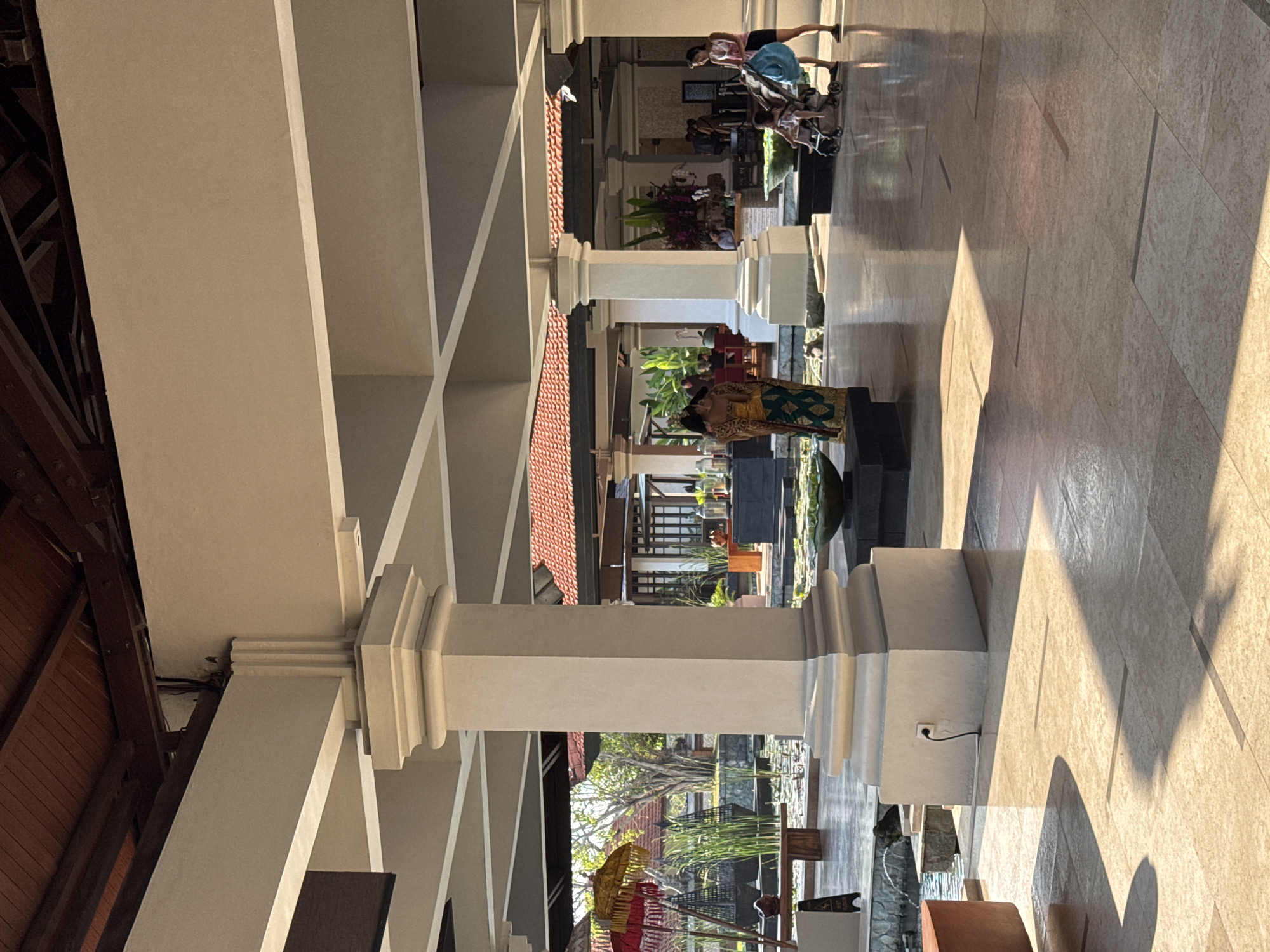 The image depicts the open-air lobby of a hotel in Kuta Selatan, Indonesia. A large, beige column supports an overhead structure with wooden beams visible above. The floor is tiled with large, light-colored tiles, and a sunlit patch with distinct shadows is visible on the right side of the frame.
In the mid-ground, two women are present. One, wearing a traditional Balinese costume, stands facing away from the camera. To the right, another woman pushes a stroller with a child in it. In the background, there is a water feature with lily pads and decorative urns. Red and yellow umbrellas are visible near the water feature, and more buildings with terracotta roofs are seen in the distance. Plants and trees are also part of the environment. The overall impression is that of a tranquil, tropical hotel lobby.
