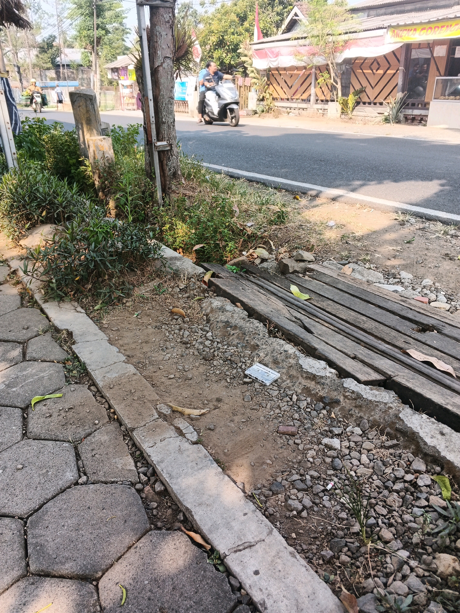 The image depicts a street scene in Ungaran, Indonesia, with a focus on the roadside. A paved sidewalk, constructed with hexagonal gray stones, runs along the left side of the frame. Adjacent to the sidewalk is a concrete curb bordering a rough, unpaved area filled with gravel, dirt, and sparse vegetation. A wooden structure, seemingly a makeshift bridge or drainage cover, is partially visible in this area, composed of several weathered wooden planks laid horizontally across a gap. Scattered debris, including fallen leaves and small rocks, are present throughout this section. Further into the scene, a busy road is visible with two individuals on motorbikes. One person, wearing a dark shirt, rides a white scooter, while another, in a yellow helmet, drives a lighter-colored motorbike. Along the road, there are buildings with awnings, suggesting commercial establishments. The overall environment appears to be a warm, sunny day in an urban or semi-urban setting.