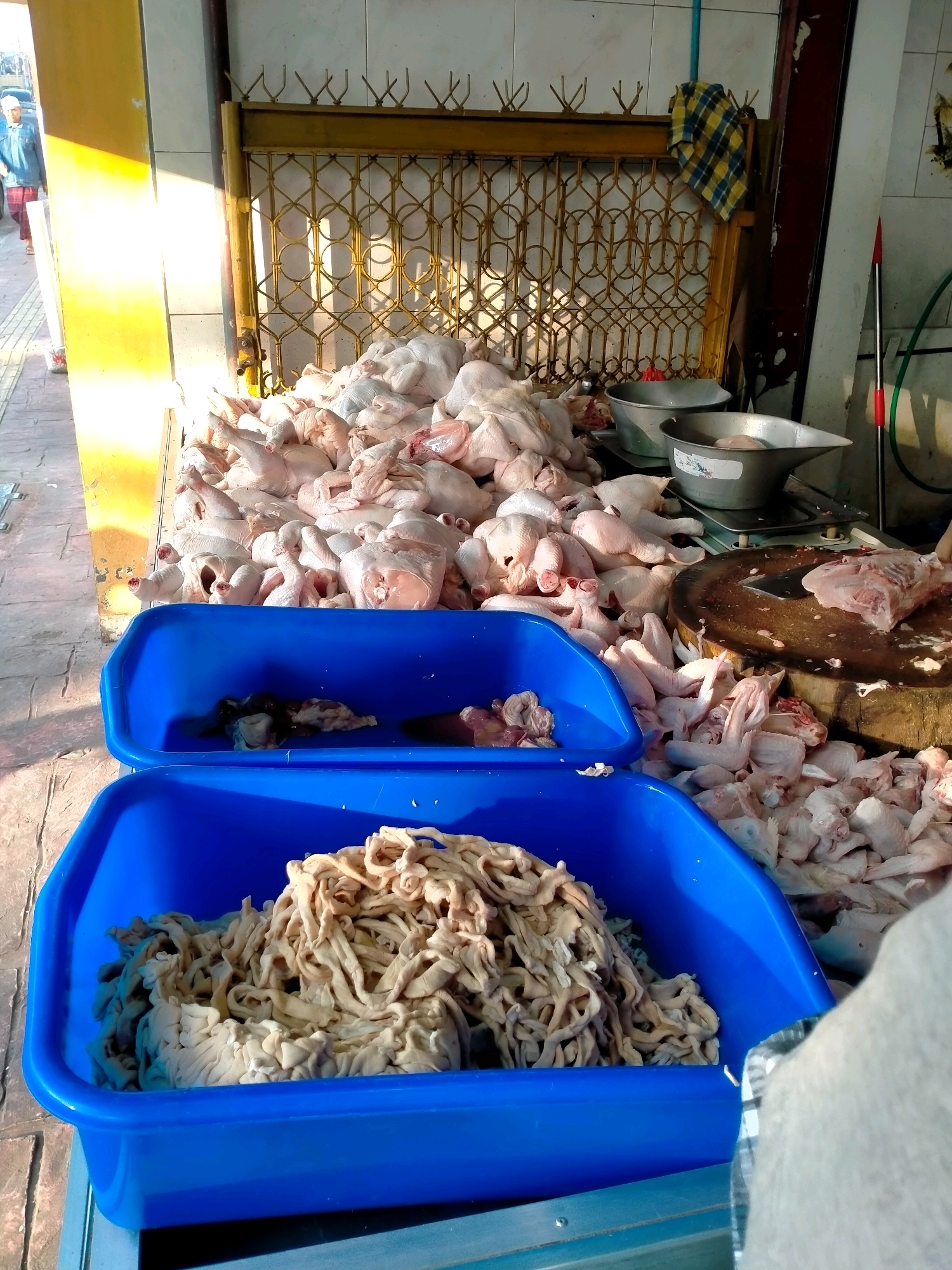 The image captures a scene at a market in the city of Batu, Indonesia, likely a butcher's stall. In the foreground, two bright blue plastic tubs are visible. The lower tub is filled with a large quantity of coiled, light brown intestines. The tub above it contains a smaller amount of darker, mixed offal. Behind these tubs, a substantial pile of whole, undressed chickens, some appearing to be cut up, are arranged on a counter. Parts of chickens, such as wings and pieces of meat, are also scattered around the counter.
On the counter, several metal bowls are present, and a large, circular wooden chopping block with a knife on it is prominently displayed, indicating ongoing butchering activities. In the background, a yellow metal grate structure is visible, possibly part of a stall or shopfront. A checkered cloth hangs on the right side of this structure. A person, partially visible and out of focus on the far left, is walking away from the camera. The lighting suggests an outdoor or semi-open environment, with sunlight casting strong shadows on the counter and the ground. The overall impression is of a busy, working market stall specializing in poultry.