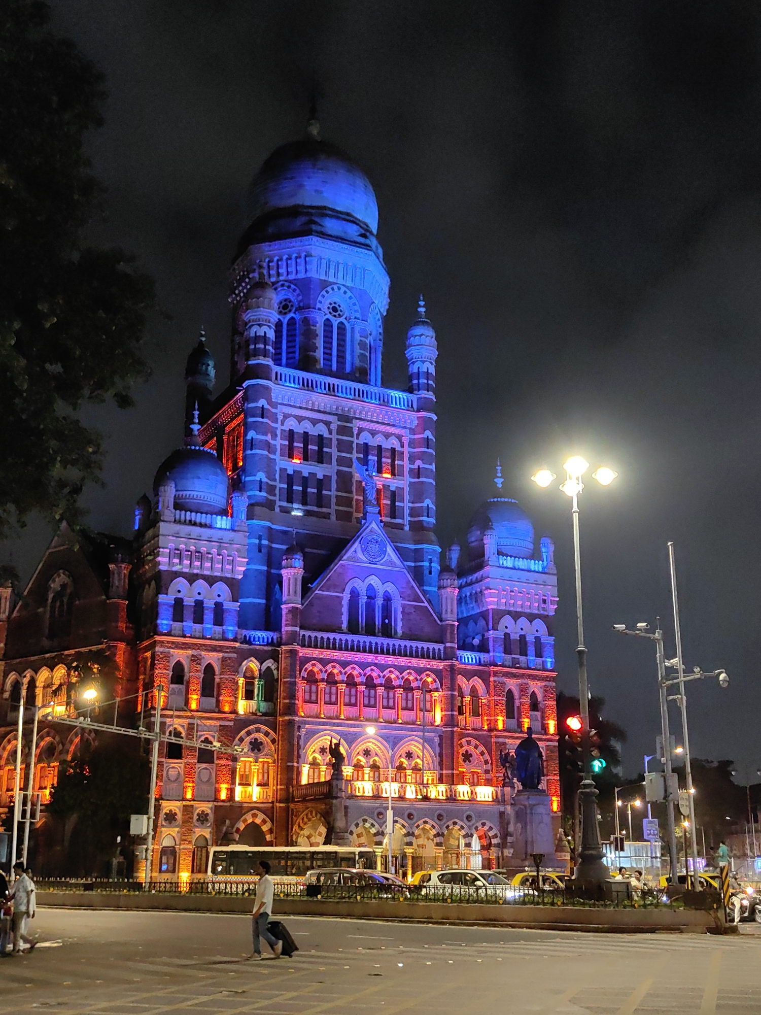 The image captures a nighttime view of the Chhatrapati Shivaji Maharaj Terminus (CSMT) in Mumbai, India. The iconic building is illuminated with vibrant blue and orange lights, highlighting its intricate Victorian Gothic architecture. A large dome and multiple spires are visible, as well as ornate windows and decorative stonework.
In the foreground, a street scene unfolds. A man is seen pulling a suitcase across a pedestrian crossing. Several cars, including a bus, are present on the road. Streetlights and traffic signals illuminate the area, casting a warm glow on the asphalt. The sky is dark and overcast, characteristic of a night in a bustling city. The overall atmosphere is one of urban activity and architectural grandeur.