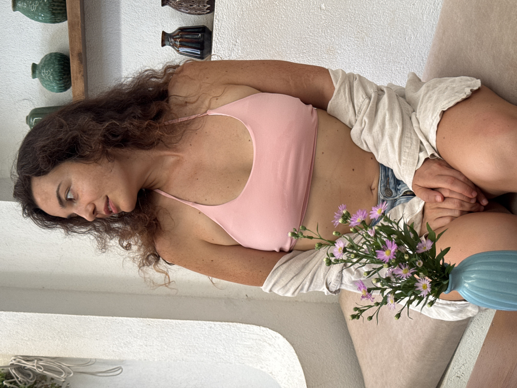 A woman with curly brown hair and freckled skin is sitting on a cushioned bench in what appears to be an outdoor cafe or seating area. She is wearing a pink halter-top bikini and a light-colored unbuttoned shirt draped over her shoulders. Her left arm is resting on her lap, and her right hand is holding a small bouquet of purple and white flowers in a blue ribbed vase. The background shows a white textured wall with a curved architectural element on the left and a wooden shelf with several decorative green and brown vases on the right. The overall setting suggests a relaxed, possibly tropical, environment, consistent with a location in Hoi An, Vietnam.