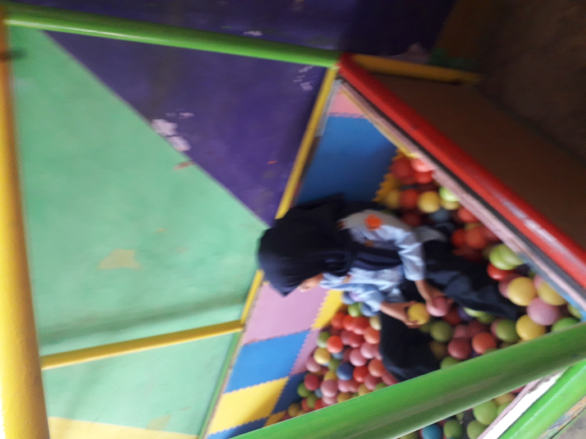 A child wearing a black hijab and a blue uniform with orange accents sits in a ball pit. The ball pit contains numerous colorful plastic balls in shades of pink, yellow, green, blue, and red. The pit is surrounded by a padded, multi-colored wall made of interlocking foam mats in yellow, blue, and pink. Above the ball pit, a structure with yellow, green, and purple-colored walls is visible. The overall scene appears to be an indoor play area, likely a playground or entertainment center, possibly located in the city of Cankuang, Indonesia.