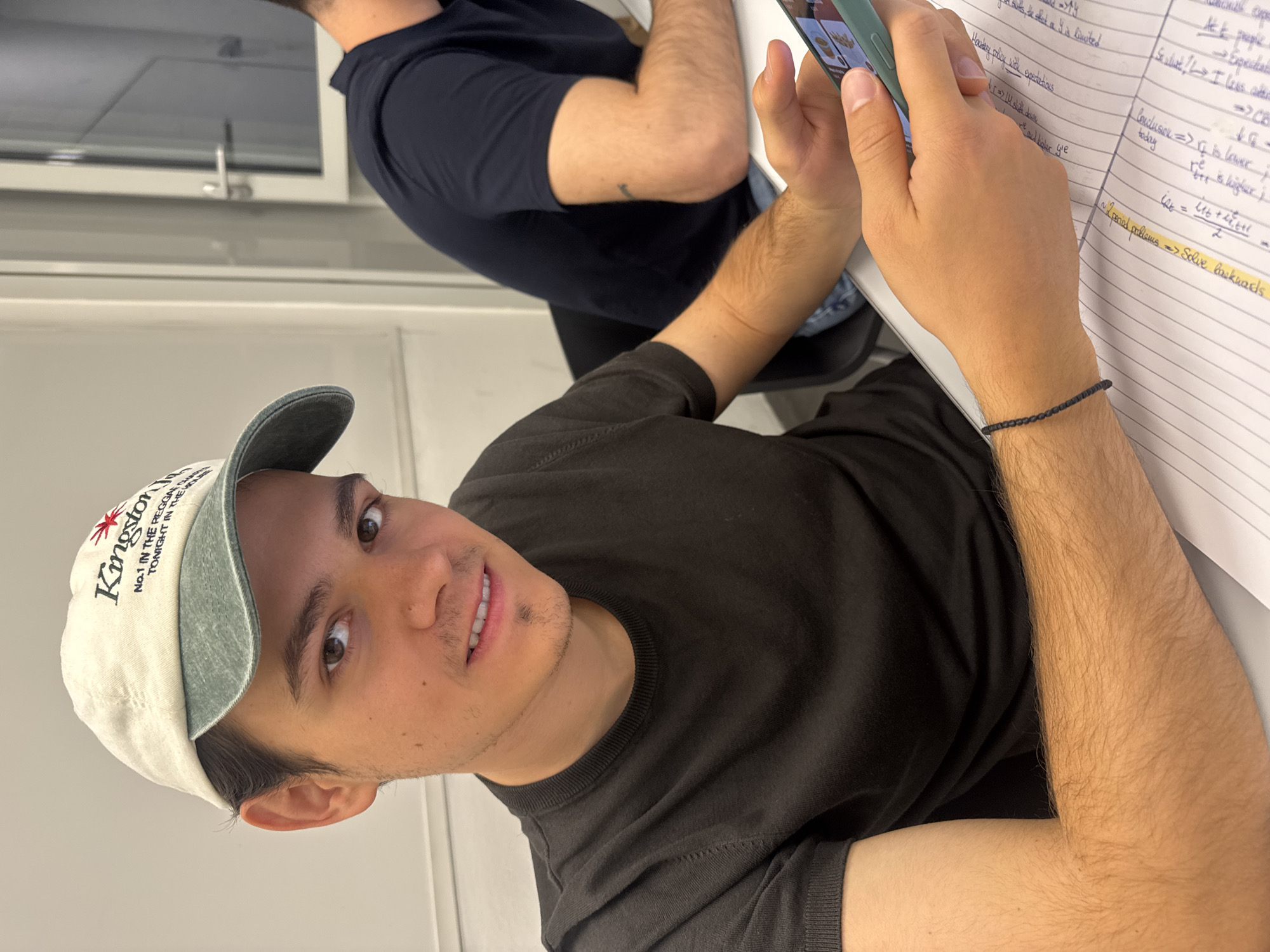 A medium shot, taken at eye-level, shows a young man wearing a faded beige baseball cap, emblazoned with "Kingston NO.1 IN THE REGGAE TONIGHT IN THE..." in black lettering with a red starburst, sitting at a desk alongside another individual. The man is holding a cell phone in his left hand. In front of him is a notebook filled with mathematical equations and notes. The immediate setting appears to be an indoor classroom or study environment with white walls. The image does not provide enough information to ascertain that this photo was taken in Milan, Italy.