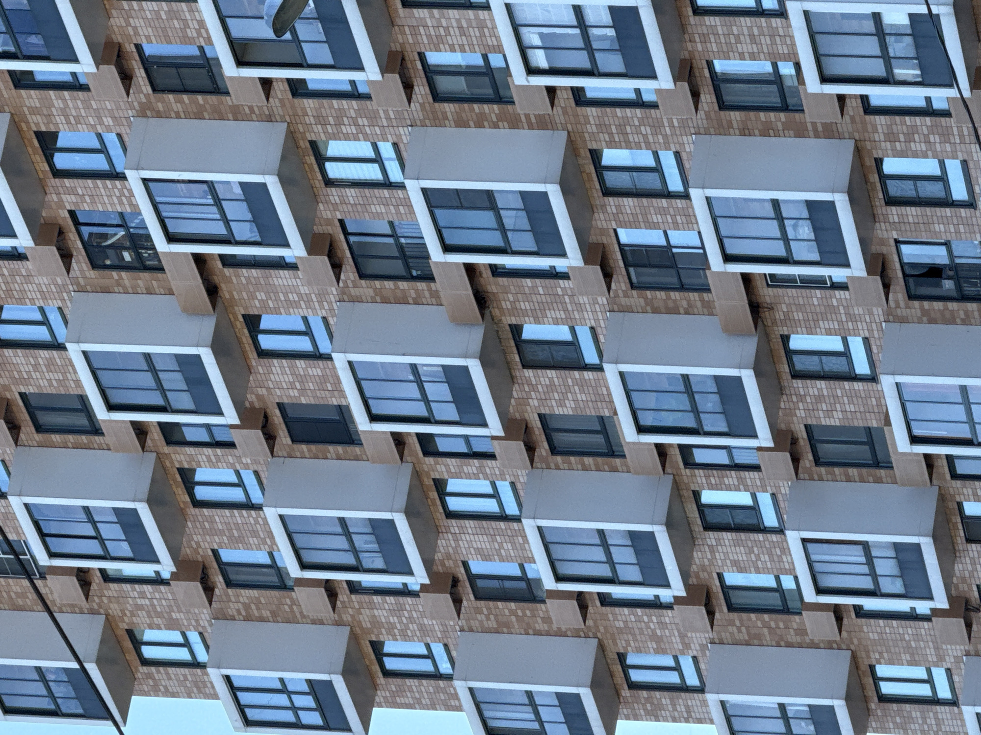 The image shows a repeating pattern of a building facade. The building has a brick exterior with protruding, angular window bays that have a grey overhang. The windows have multiple panes with black frames, and reflect a blue sky. The repetitive nature of the image suggests it may be an artistic rendering or a pattern created digitally using a photograph of the building's facade. The city of Saint Petersburg, Russia, is not discernible from the provided image.
