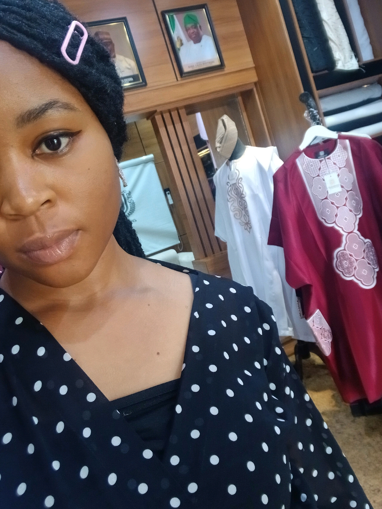 A medium shot is taken from a low angle, capturing a woman with dark hair styled in braids and a light pink clip in her hair from the chest up. She is wearing a black dress with white polka dots. Her face is turned to the left, with her eyes looking directly at the camera. She has on subtle makeup. In the background, on a wooden shelf, are framed pictures of men in official attire, and two mannequins dressed in traditional African clothing. One mannequin wears a white robe with intricate gold embroidery, and the other wears a maroon robe with white floral patterns. Rolls of fabric are stacked on shelves in the upper right corner of the image. The overall setting appears to be a store.